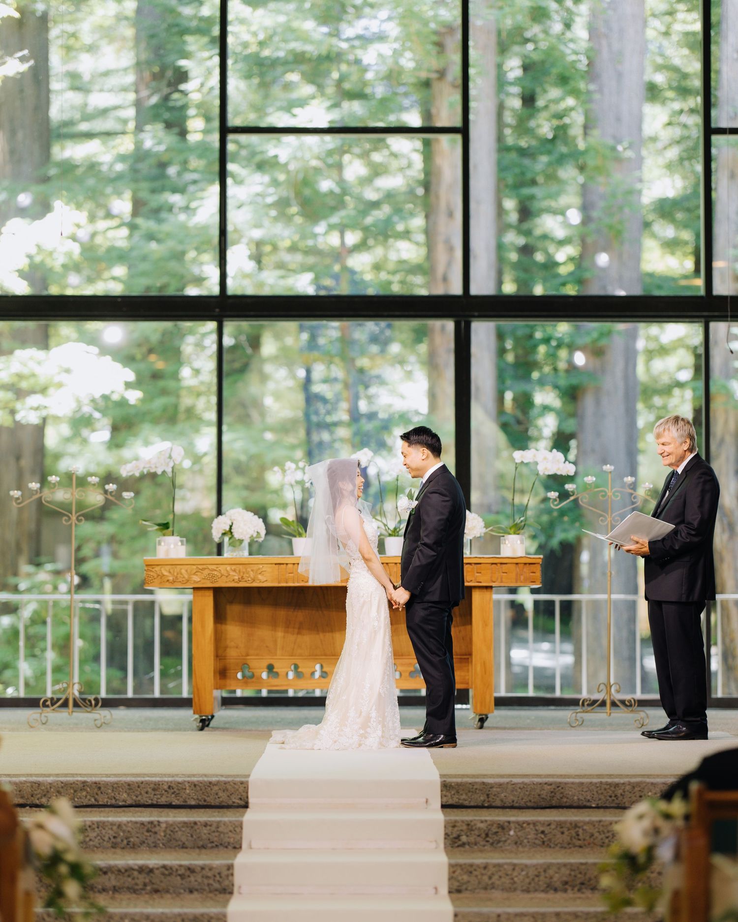 Bride and groom exchange vows at altar in elegant chapel with floor-to-ceiling windows overlooking tall trees in Bay Area wedding.