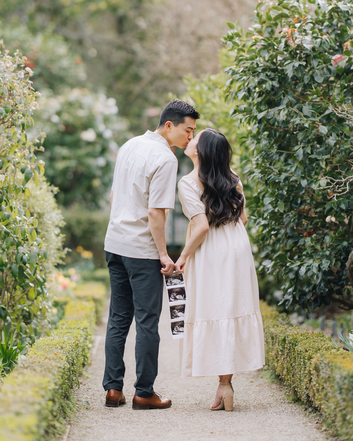 Couple sharing a kiss while holding ultrasound photos on a lush garden pathway lined with greenery.