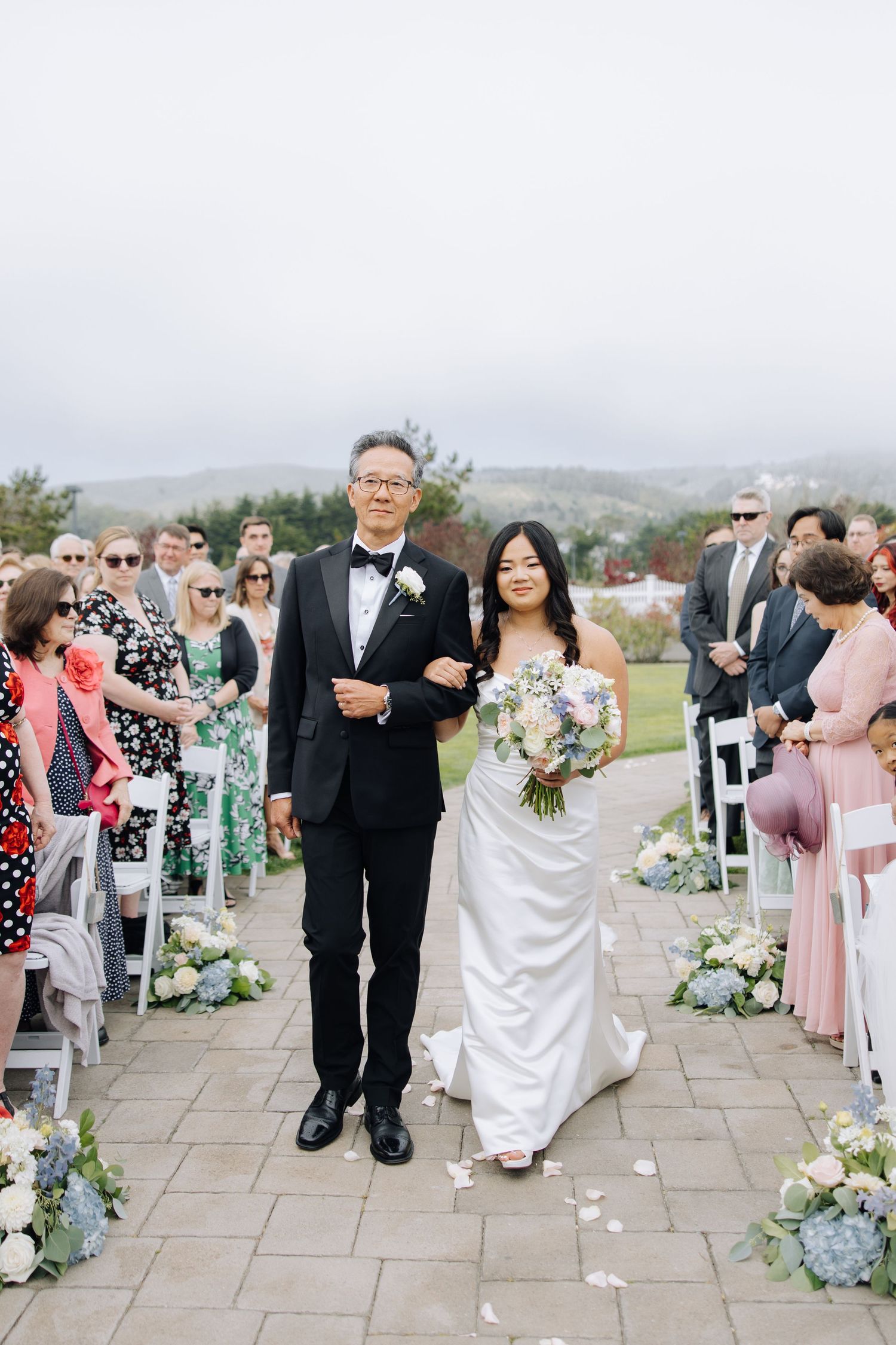 Bride walking down the aisle with her father at an outdoor wedding ceremony on a cloudy day in Half Moon Bay venue.