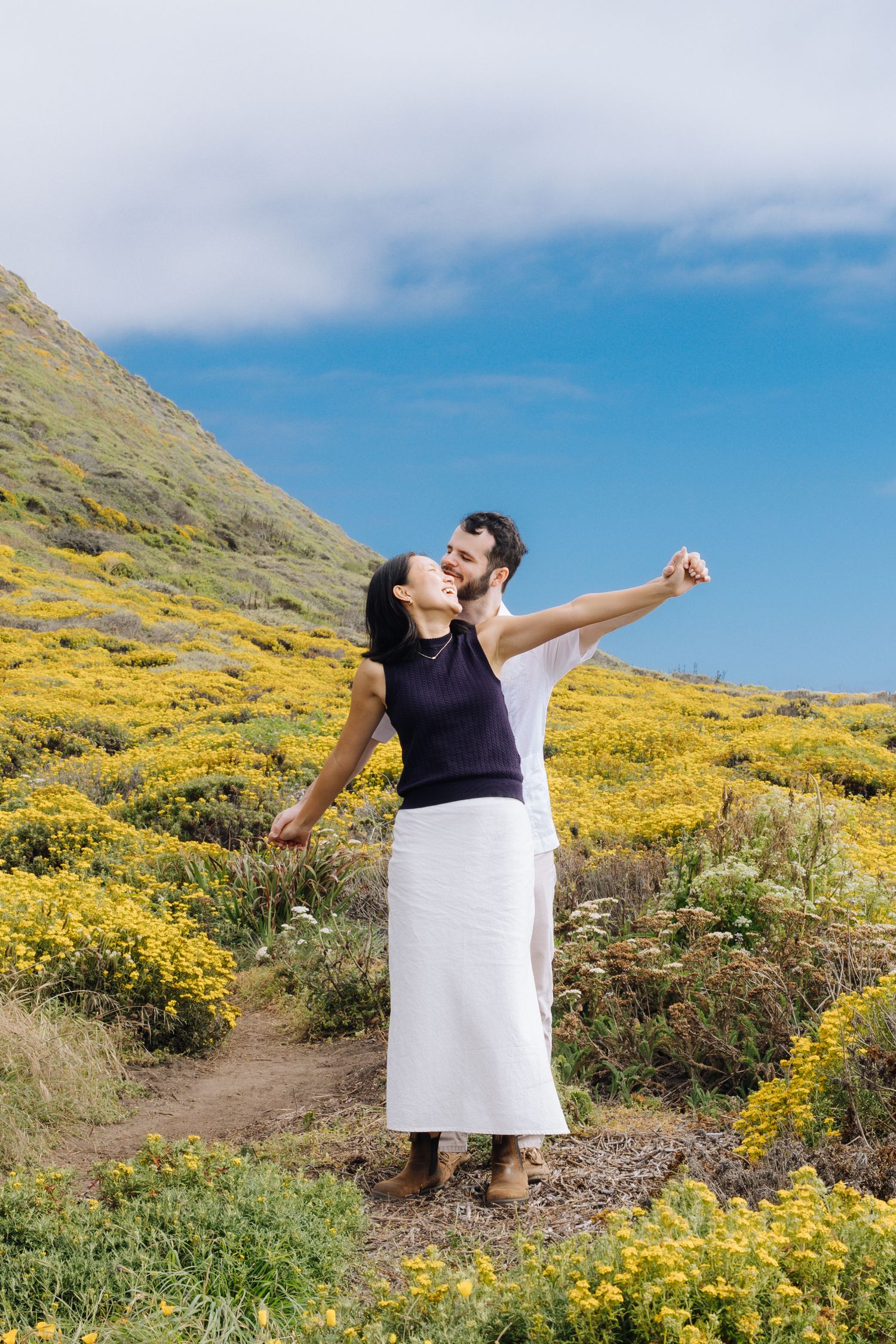 Joyful couple embracing on a hillside covered in yellow wildflowers under a blue sky during surprise proposal in Carmel.
