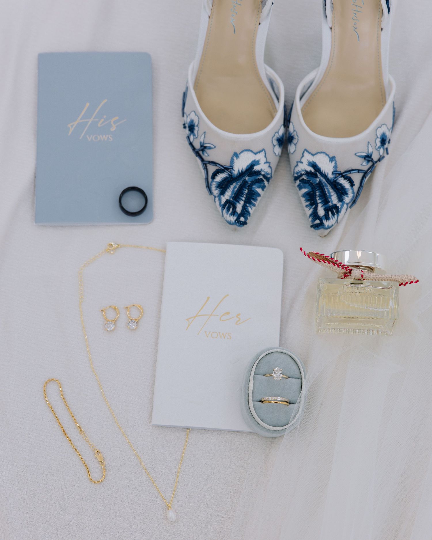 Flat lay of wedding details including blue floral heels, vow books, diamond ring, gold jewelry, and perfume.