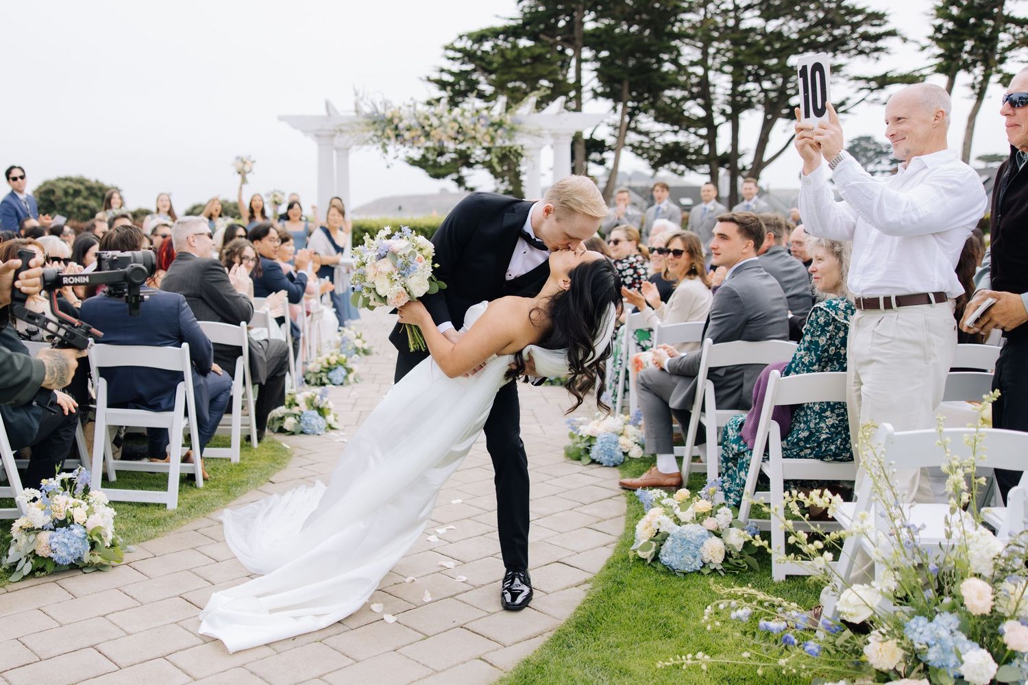 Groom dipping bride at outdoor wedding ceremony aisle lined with blue and white flowers in Half Moon Bay.