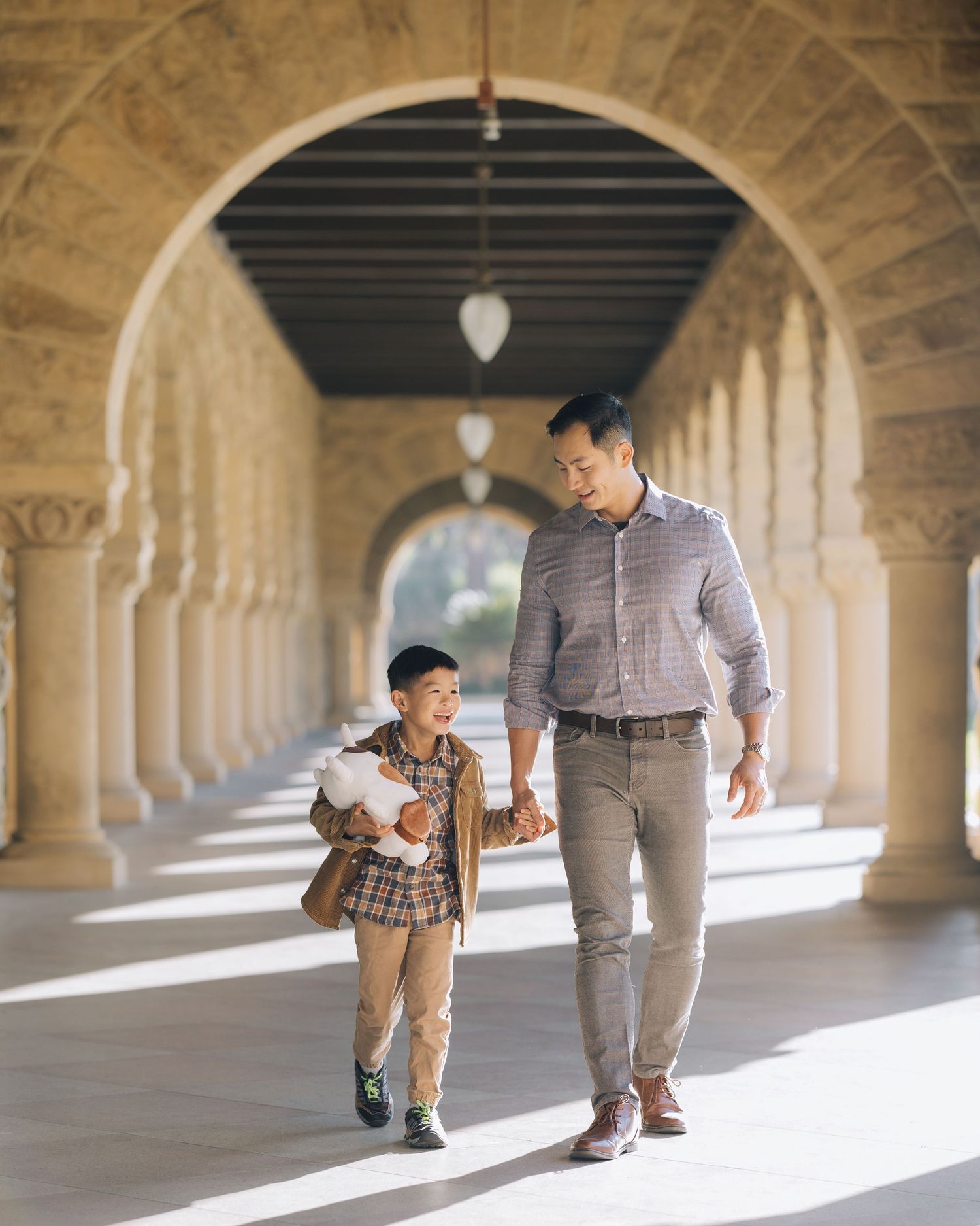 Father and son walking hand in hand through sunlit stone archway corridor at Stanford.