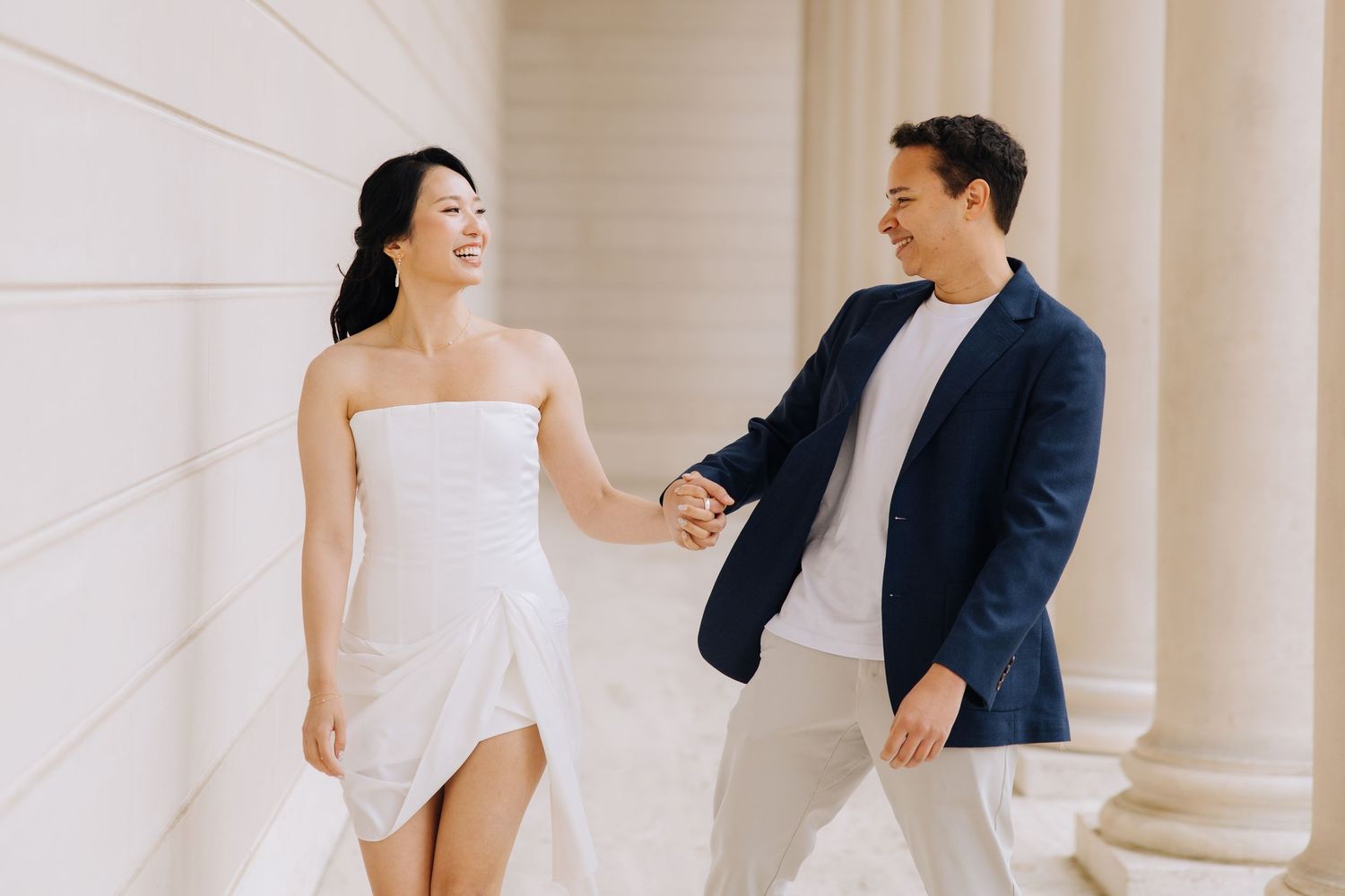 Couple holds hands and smiles walking between white columns during engagement photo session. Engagement photoshoot in Legion of Honor.