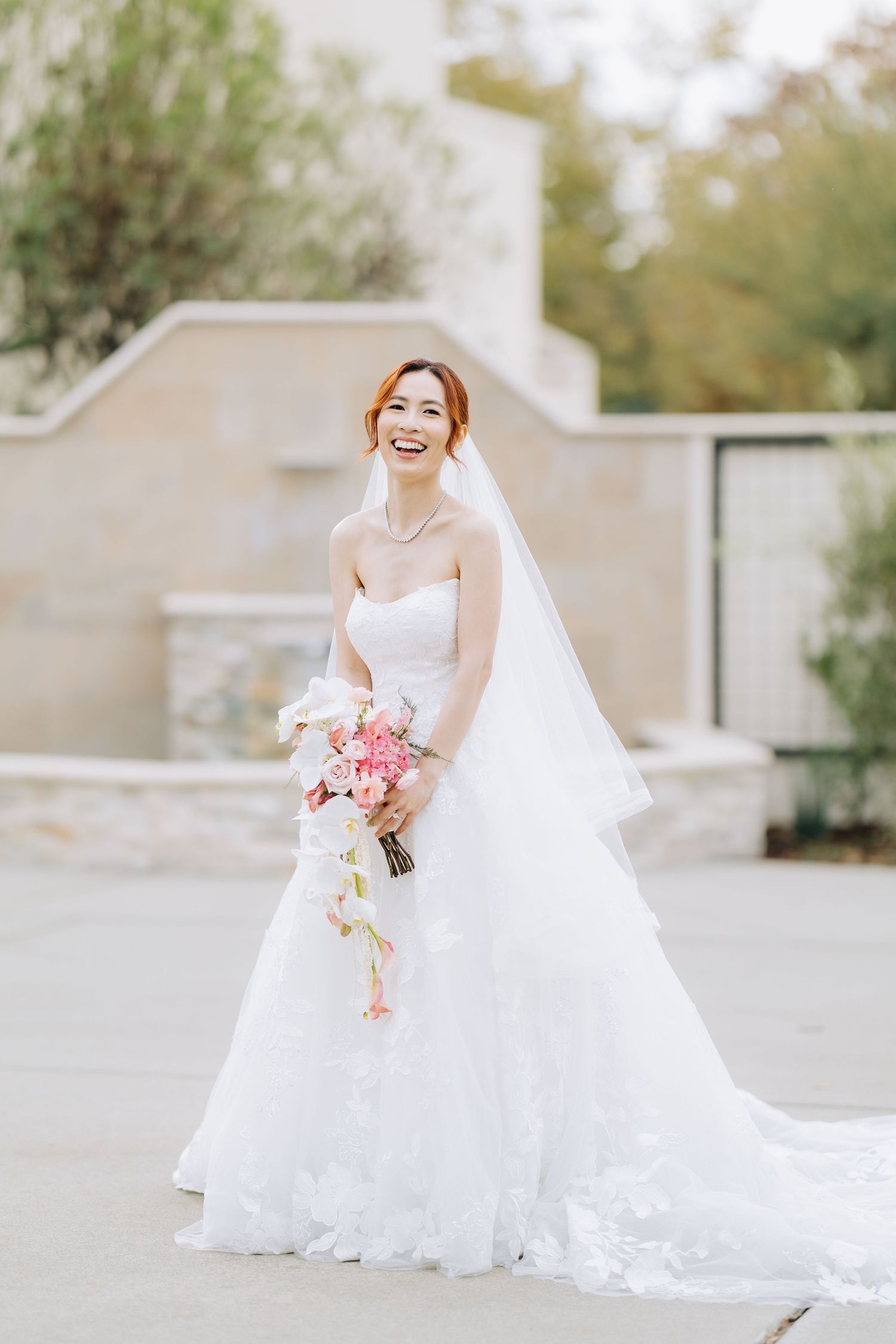 Laughing Asian bride in strapless white layered wedding gown holding pink cascading bouquet outdoors.