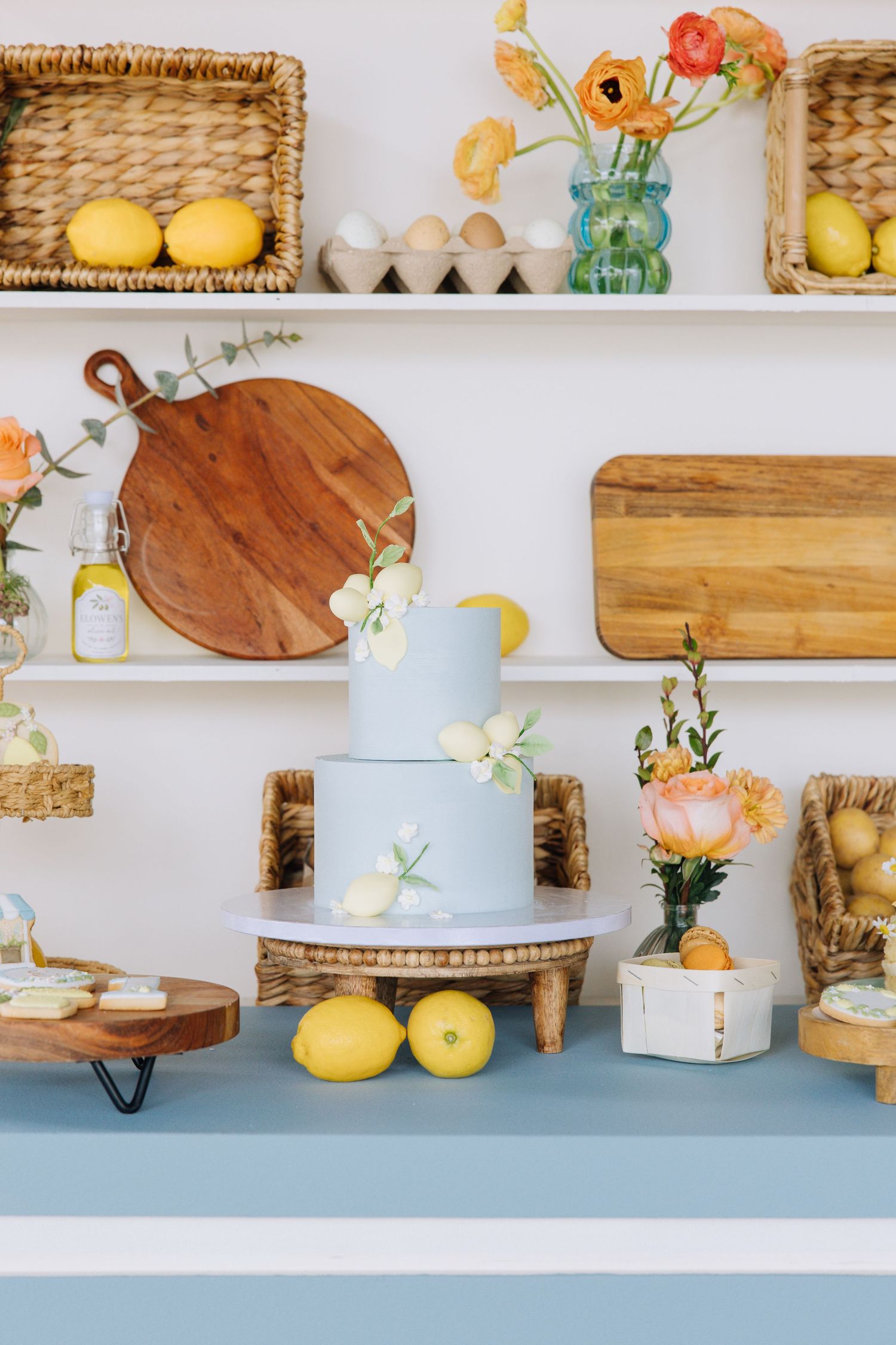Light blue two-tier cake with lemons and florals on a dessert table with wicker baskets and wooden cutting boards at baby's first birthday.
