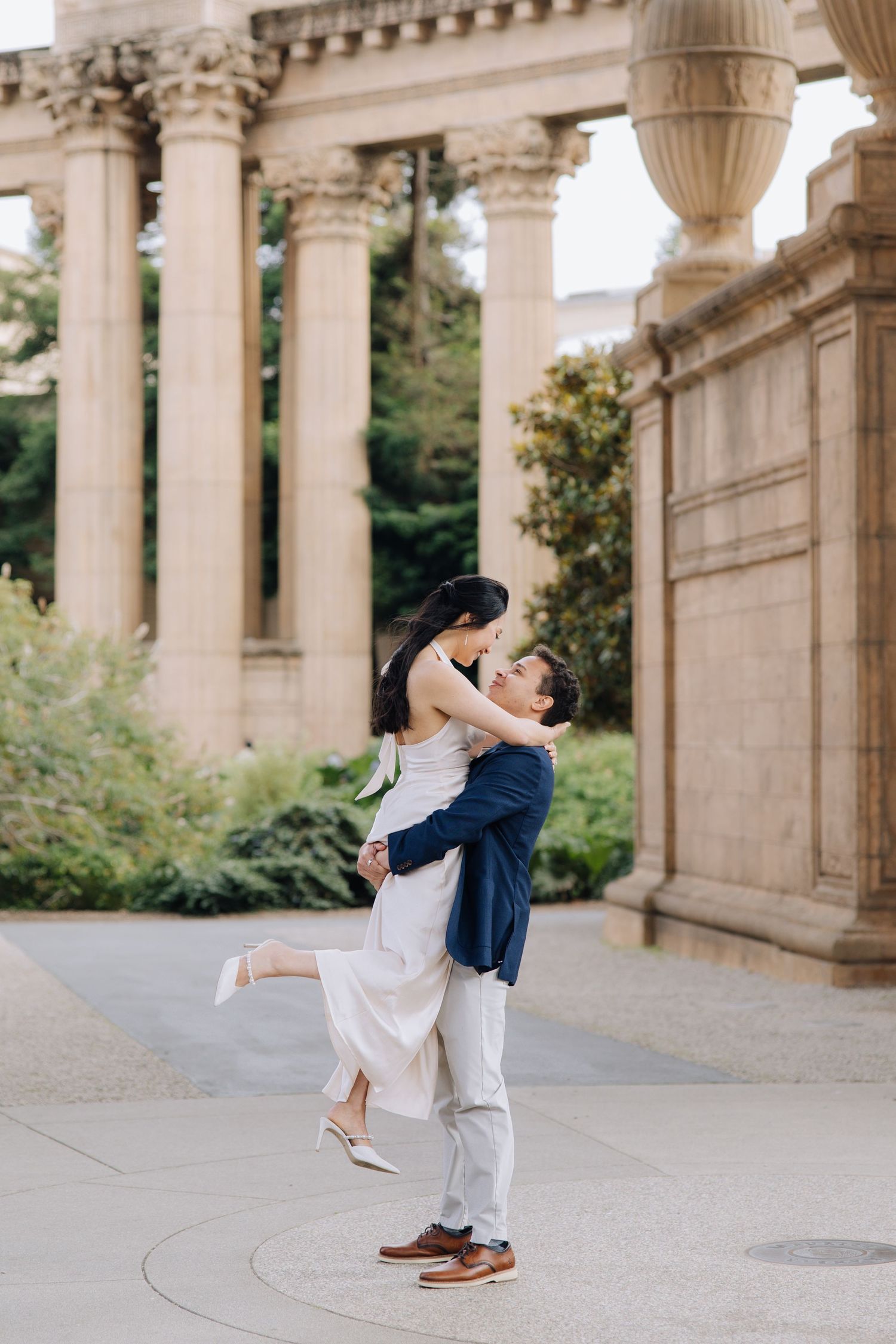 Man in navy blazer lifting woman in white dress during engagement photoshoot at Palace of Fine Arts.
