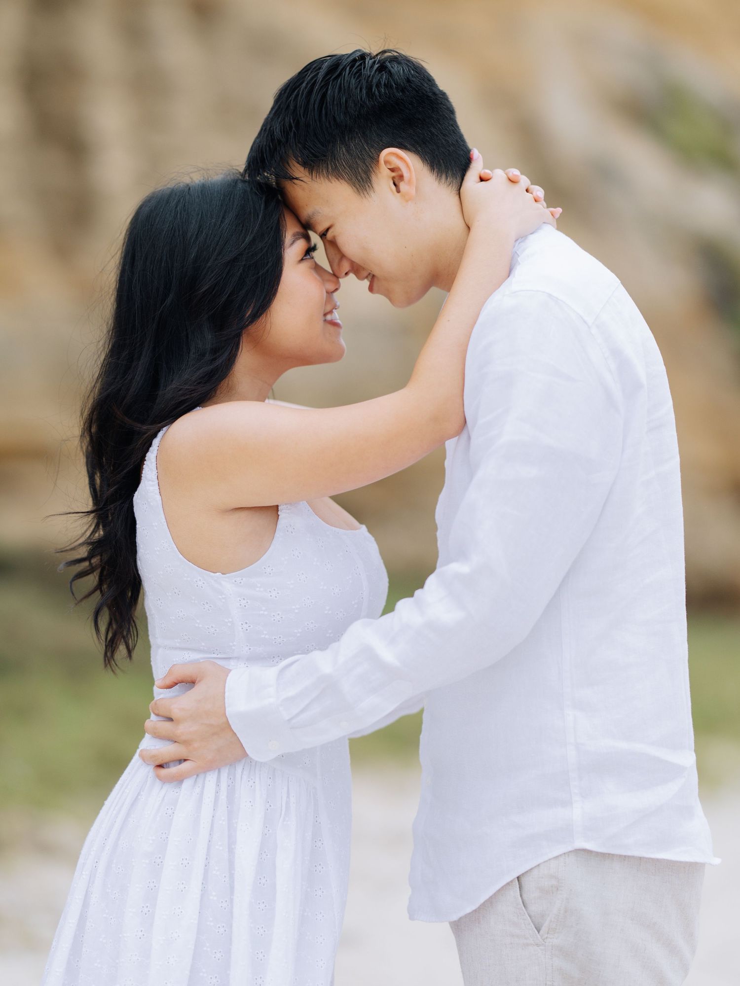 Intimate couple in matching white outfits touching foreheads tenderly at a sandy beach during Half Moon Bay beach engagement photoshoot.