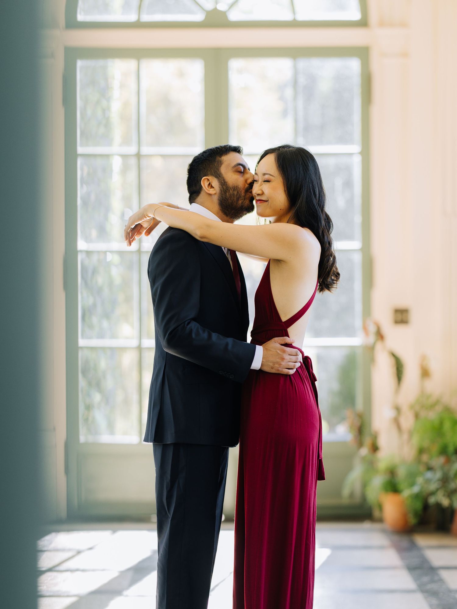 Couple sharing a romantic kiss and embrace in a sunlit indoor venue with large windows during Filoli engagement photoshoot.