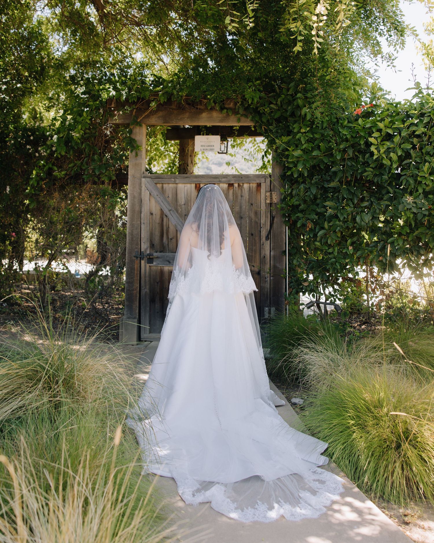 Bride in white ball gown with long veil stands before a rustic wooden garden gate surrounded by greenery at wedding in Carmel, CA.