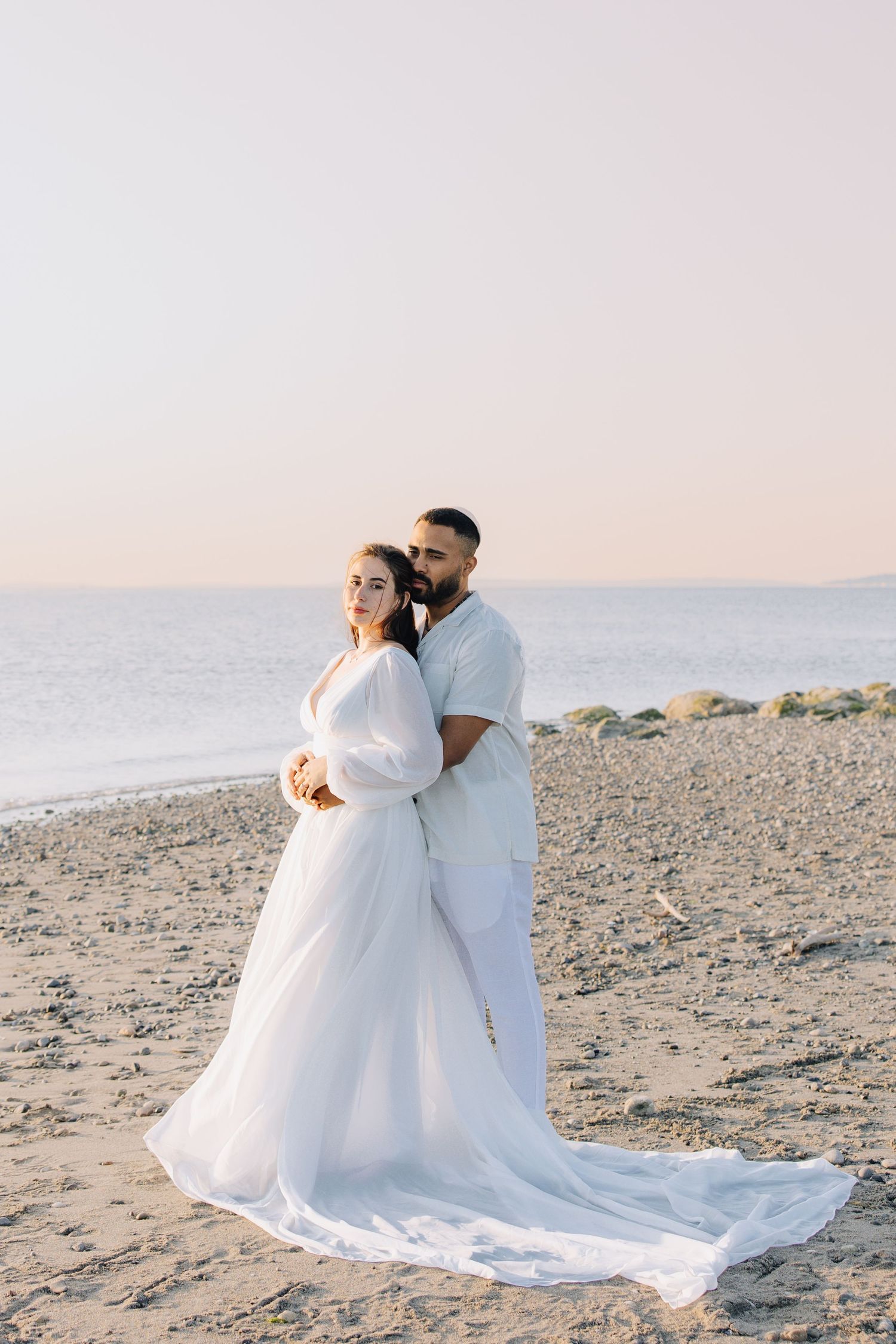 Romantic couple embracing on pebble beach at sunset, bride in flowing white gown with long train during beach engagement photoshoot.