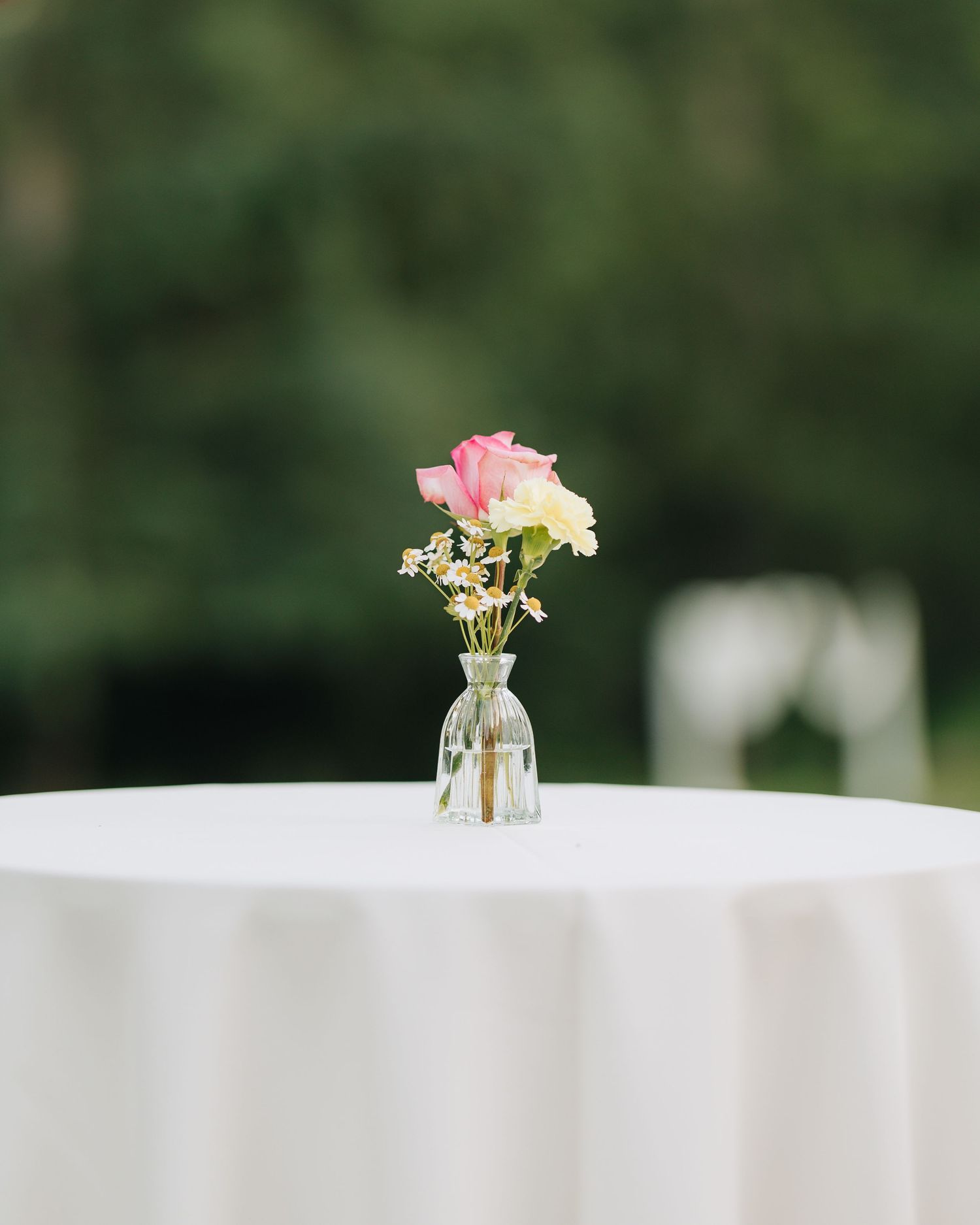 Single pink rose and white flowers in small glass bud vase on white linen cocktail table outdoors.
