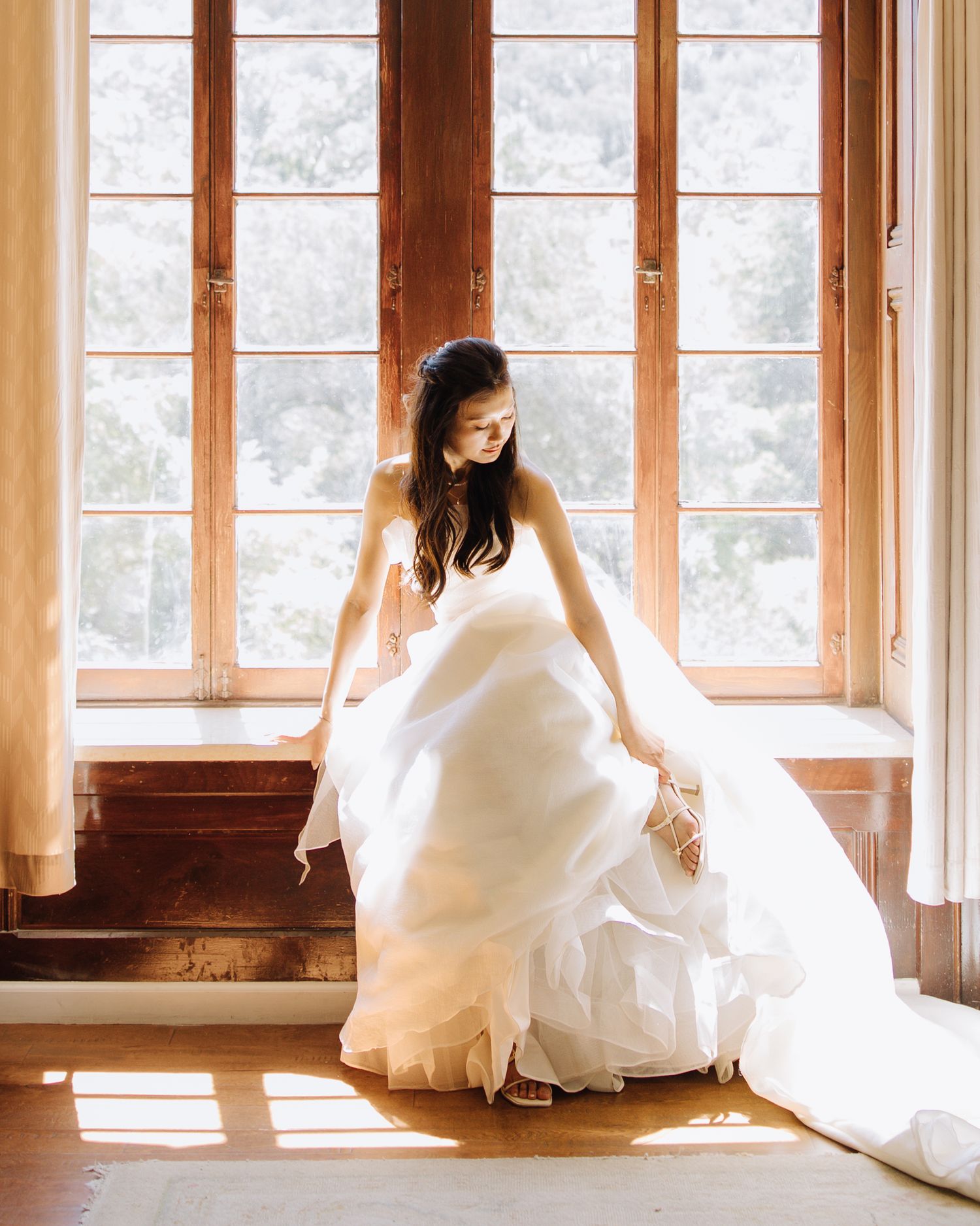 Bride in flowing white ballgown sits by large wooden French windows in soft natural light in Elliston Vineyards in Sunol, California.