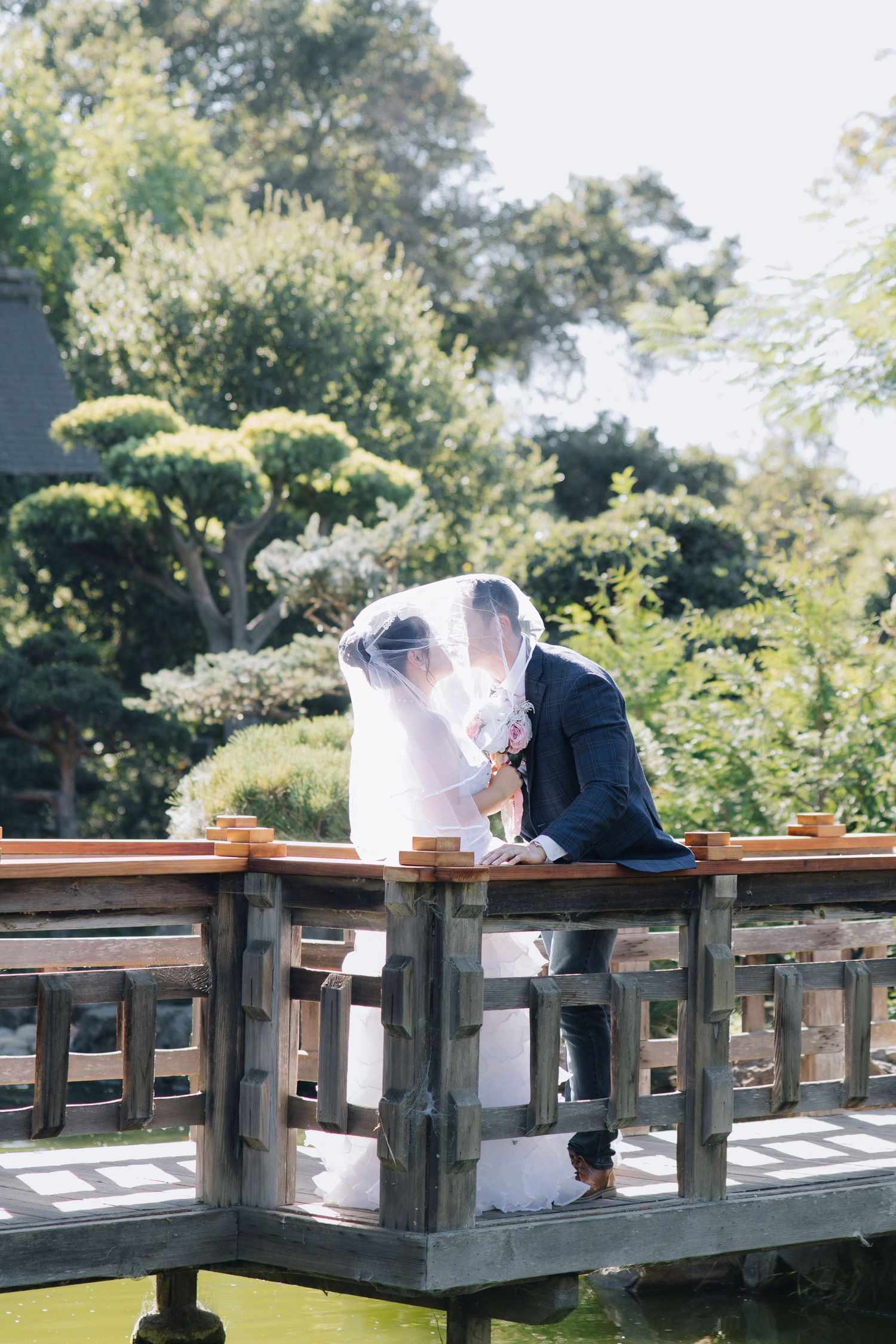 Bride and groom sharing a romantic kiss under a veil on a wooden bridge in a Japanese garden.