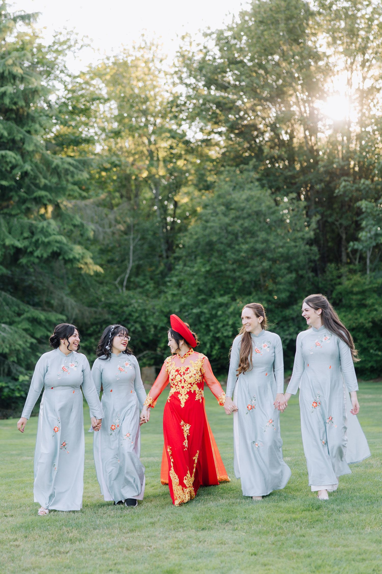 Bride in red ao dai walks with bridesmaids in light blue ao dai gowns across green lawn at golden hour.