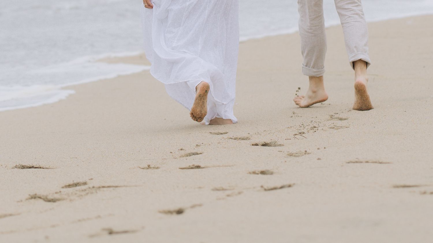 Barefoot couple walking on sandy beach leaving footprints, wearing white outfits near ocean waves during beach engagement photoshoot.