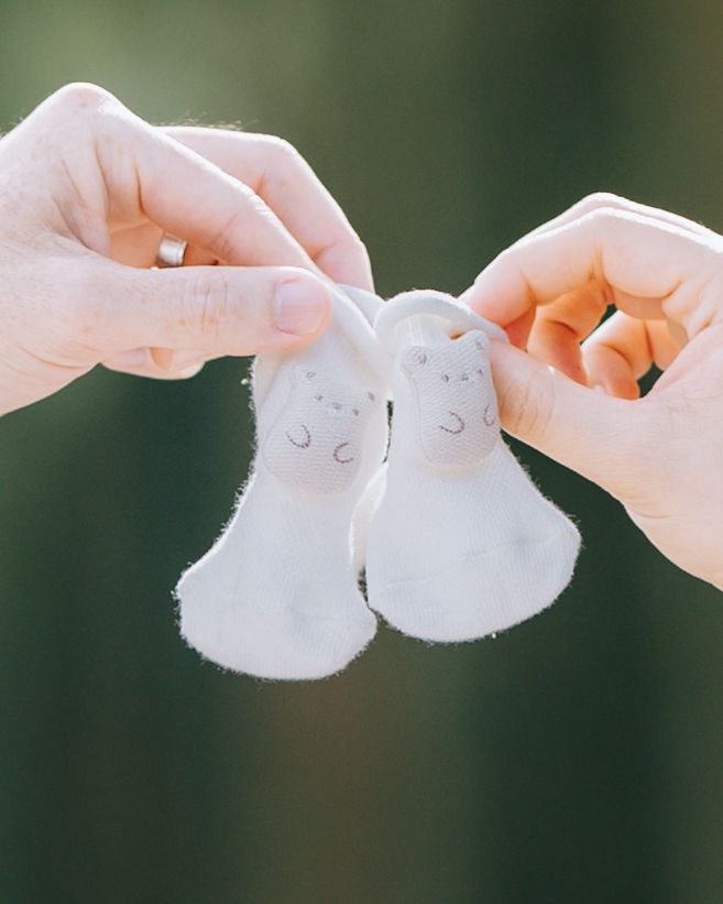 Two hands holding tiny white baby booties against soft green background in Filoli maternity photoshoot.