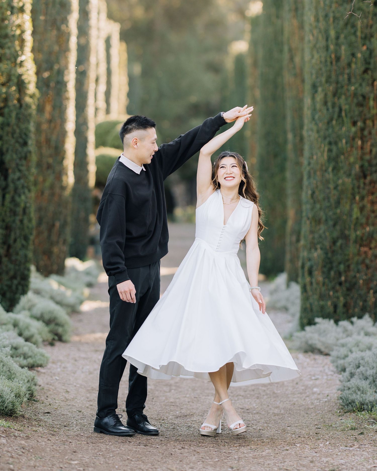 Joyful couple dancing along a cypress-lined garden path, woman twirling in white dress during Filoli engagement photoshoot.