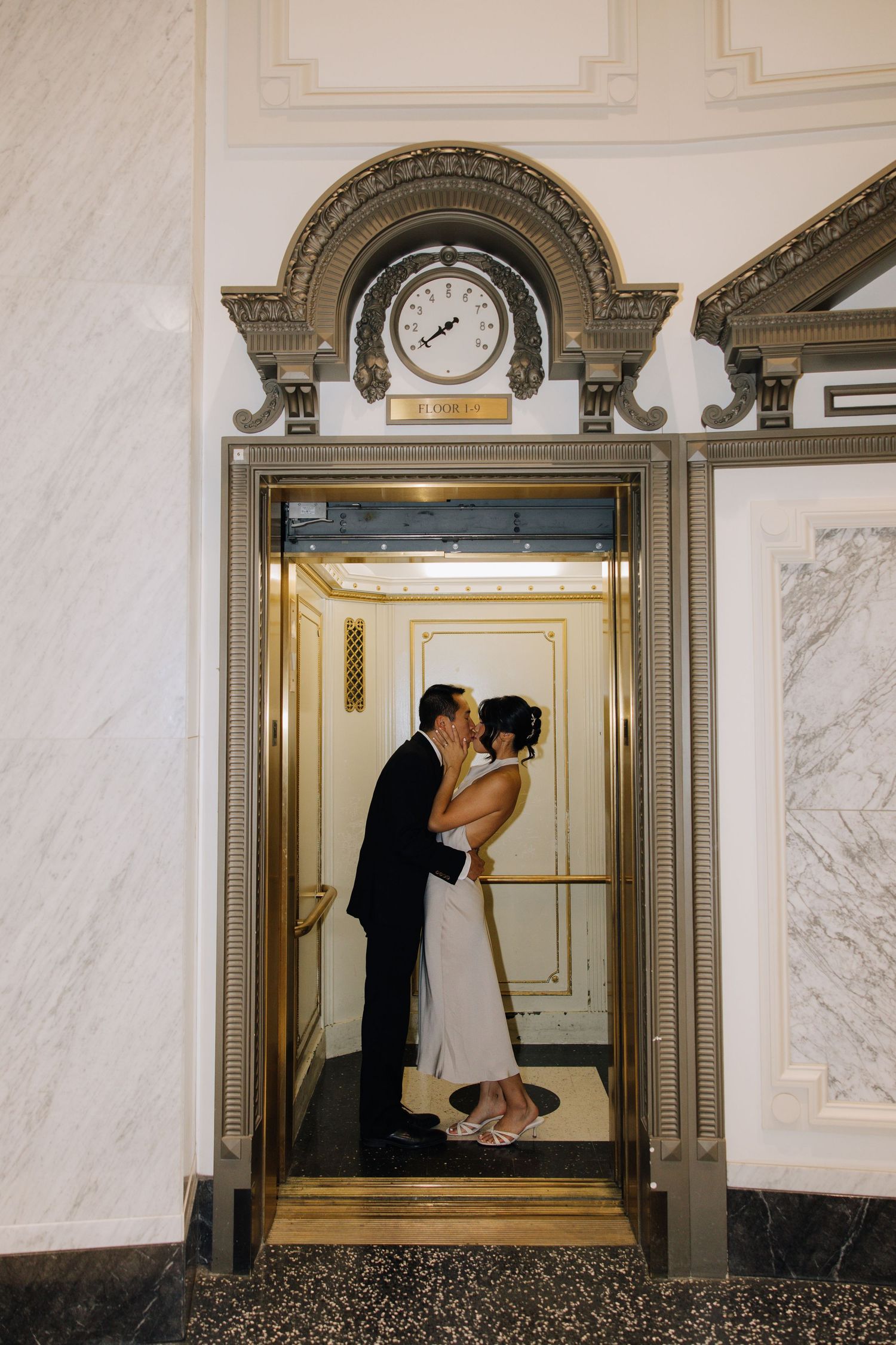 Bride and groom share a romantic kiss inside an ornate gold elevator with a decorative clock above the doors during city hall wedding.