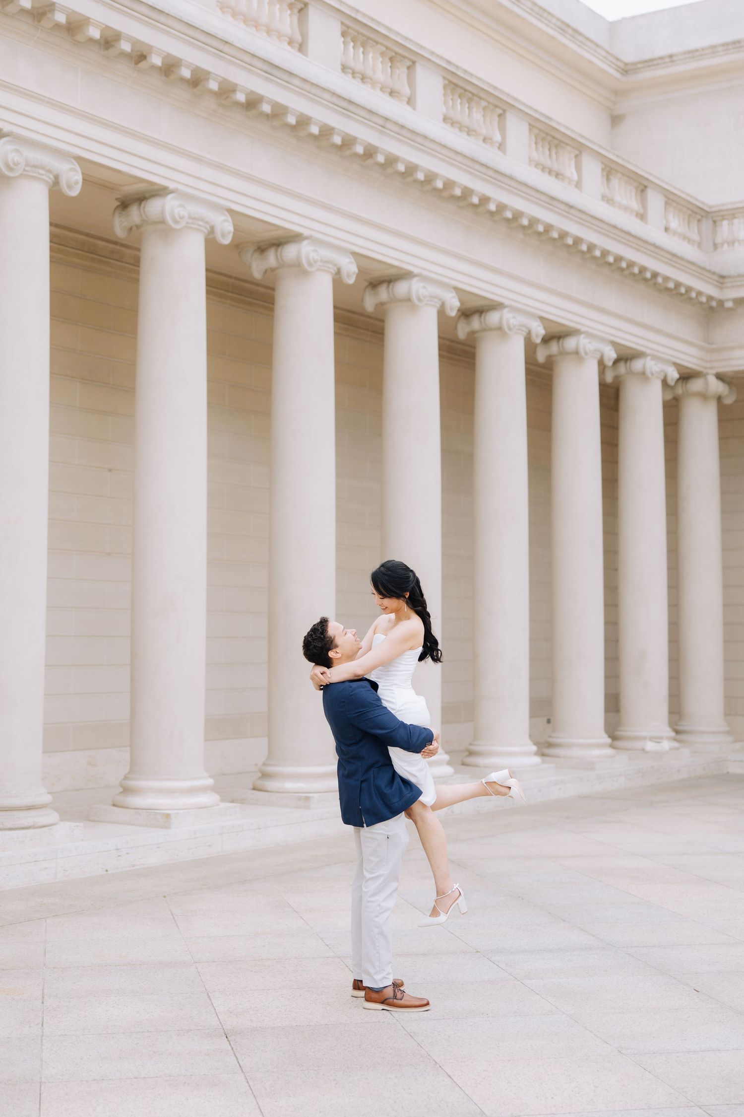 Man in navy suit lifting woman in white dress outdoors near classical stone columns during engagement photoshoot at 
Legion of Honor museum.