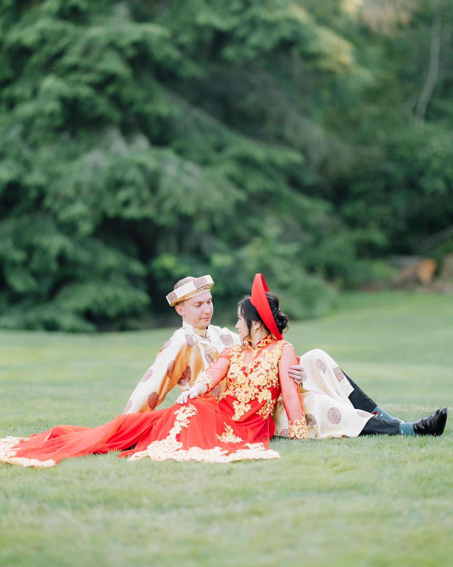 Couple in traditional wedding attire sitting on green grass, bride in red Chinese qipao, groom in white South Asian sherwani.