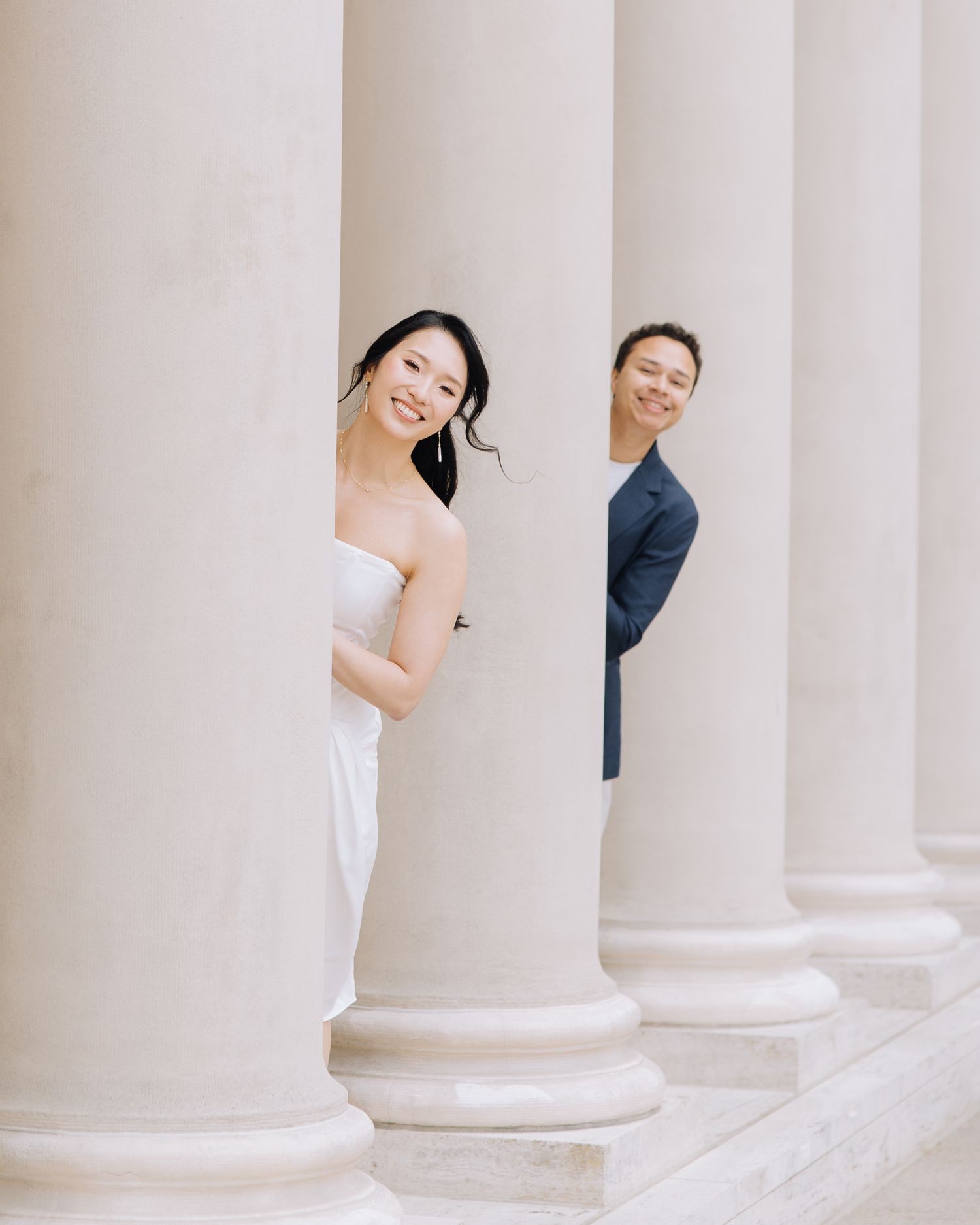 Couple peeking playfully between white columns during engagement photoshoot at Legion of Honor museum in SF.