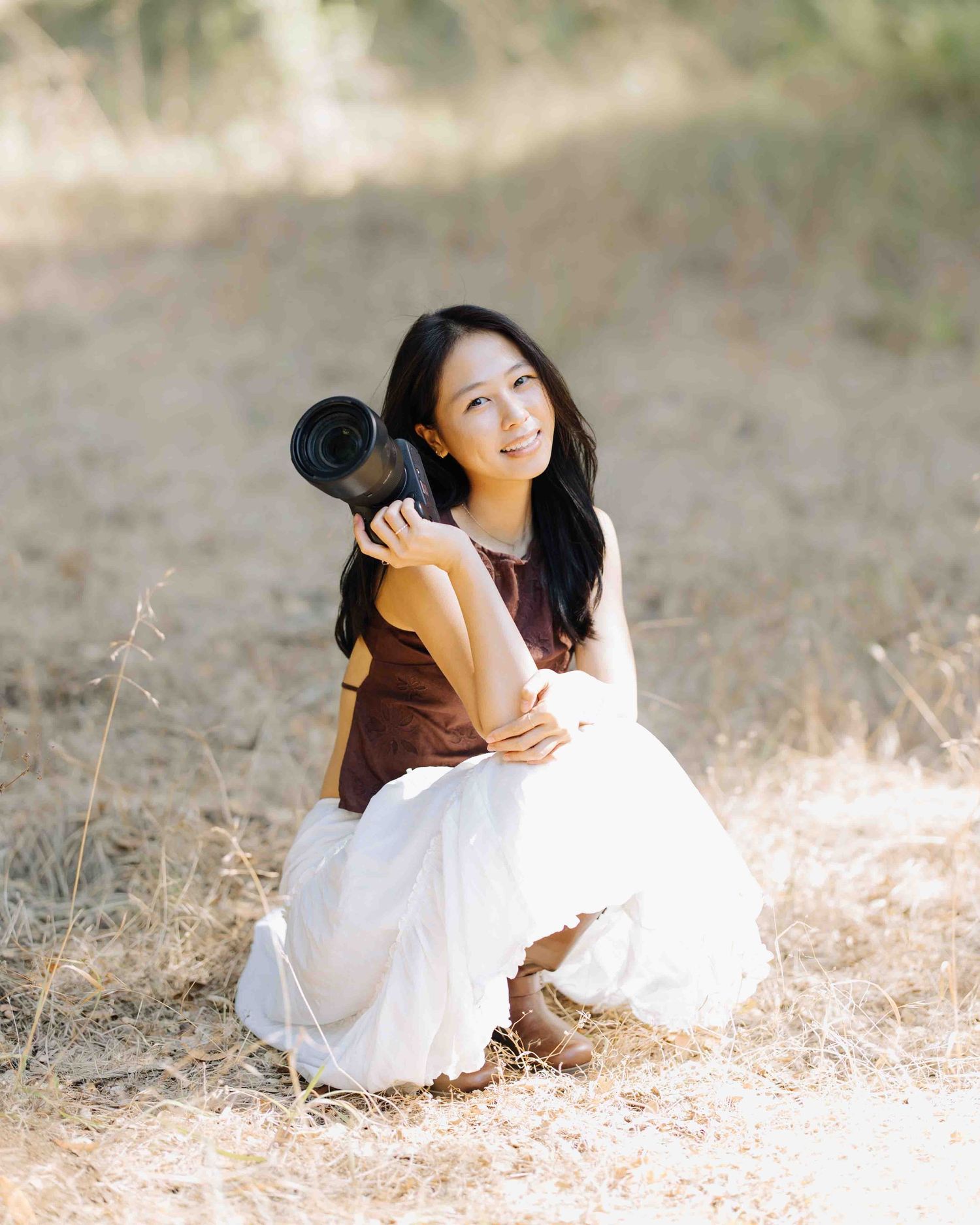Young woman photographer crouches outdoors holding a camera, smiling in natural dry grass setting.