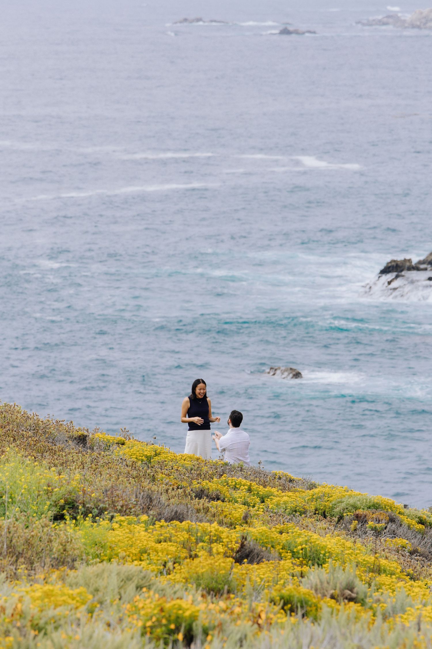 Couple standing on a coastal cliffside with yellow wildflowers and ocean waves below during surprise proposal in Carmel.