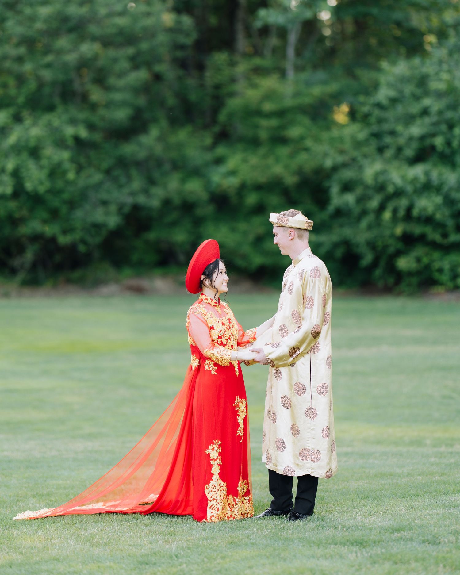 Bride in traditional Vietnamese red ao dai and groom in cream ceremonial attire hold hands on green lawn.