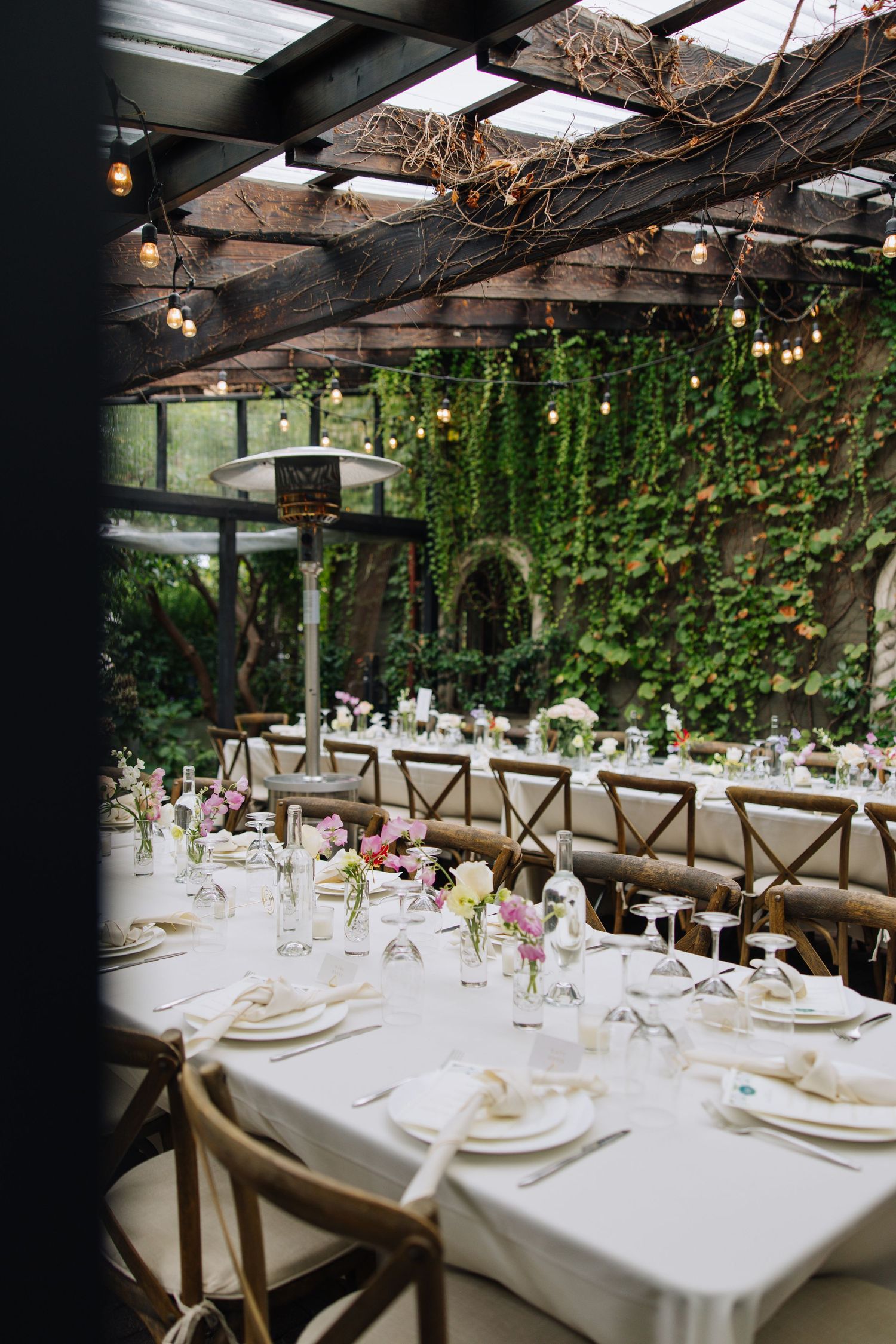 Wedding reception tables with white linens and pink floral arrangements under string lights in ivy-covered venue in Bay Area.