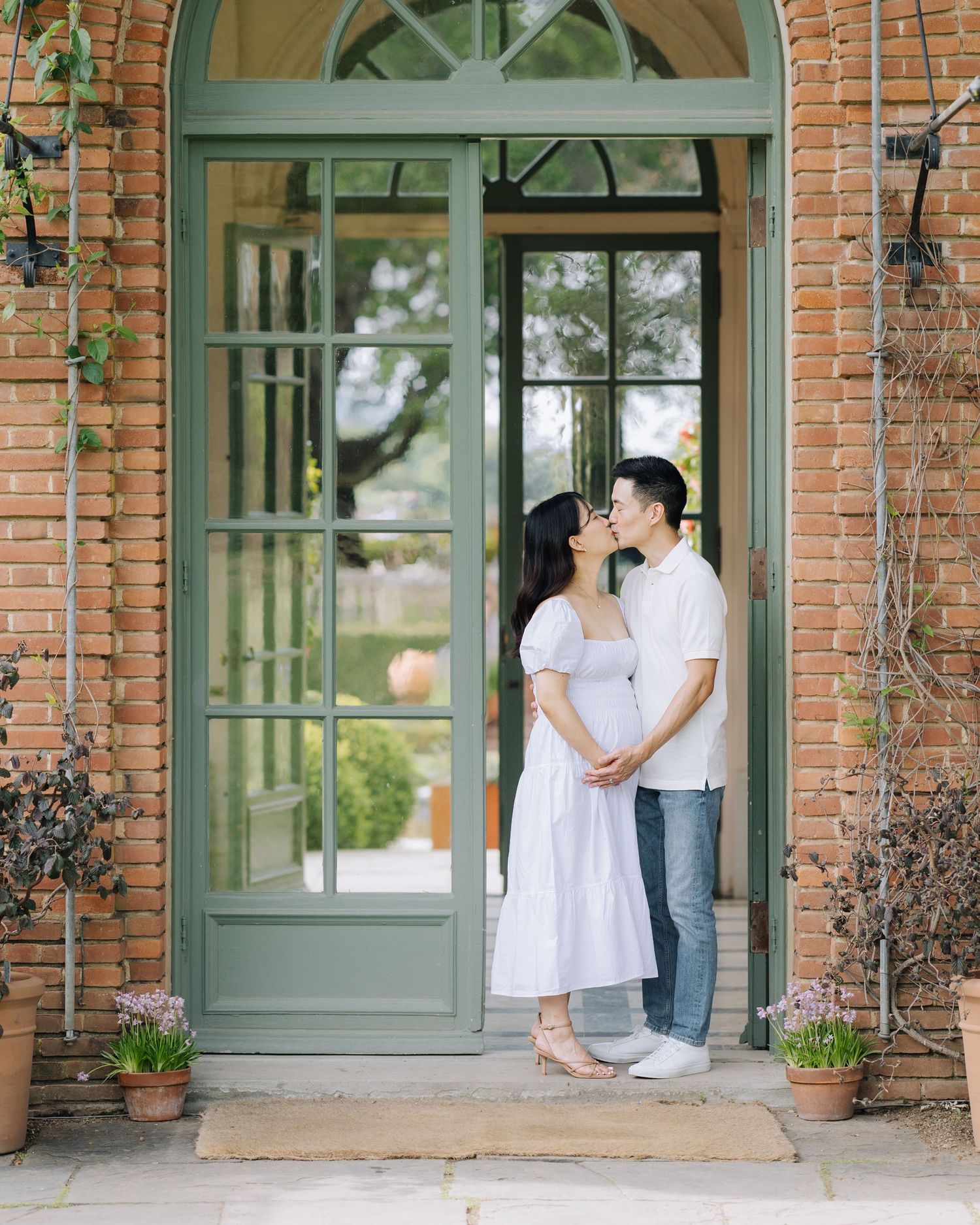 Expecting couple sharing a kiss in doorway of brick building with green French doors at Filoli Woodside maternity photoshoot.