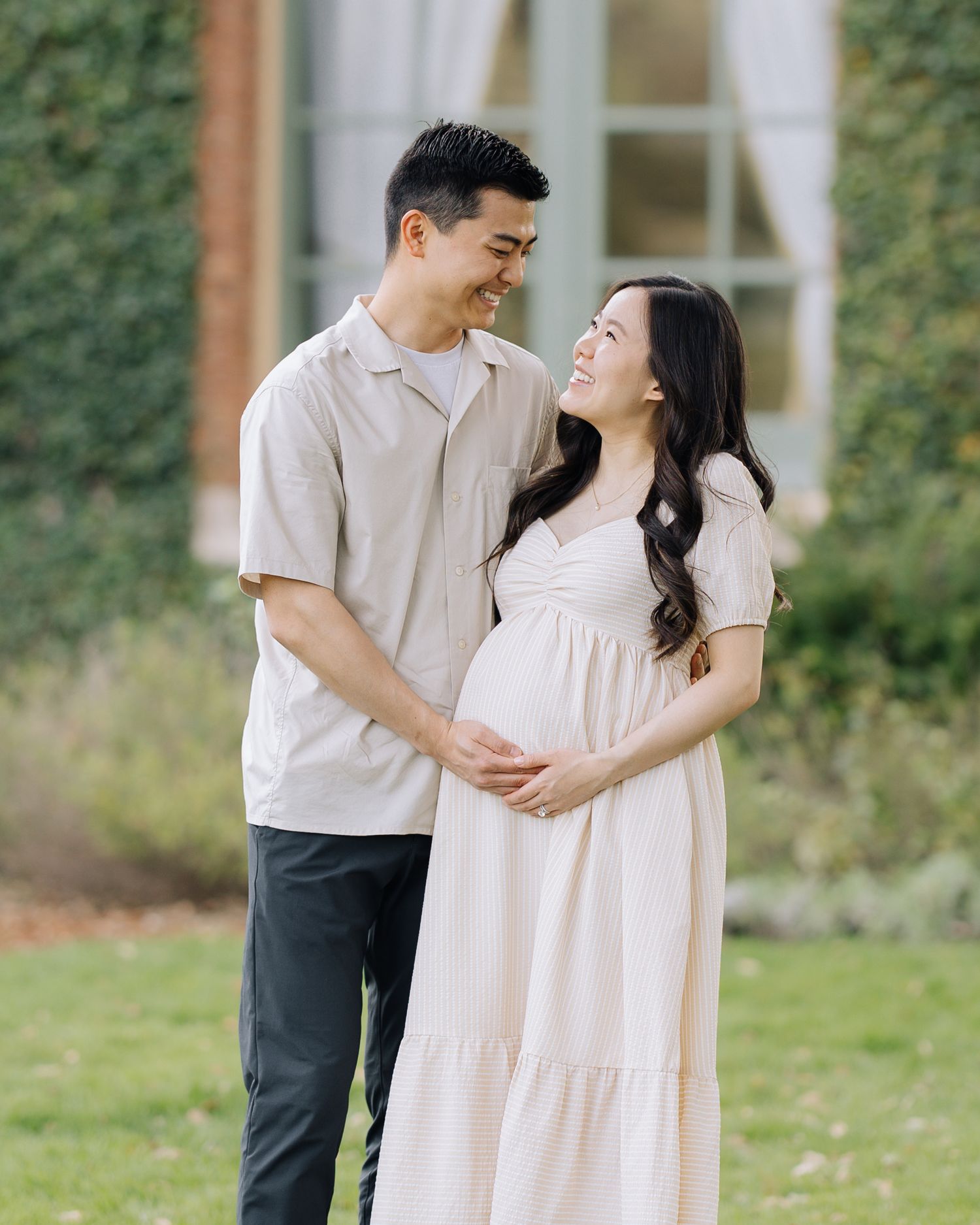 Smiling expecting couple in neutral tones embracing outdoors in a lush green garden setting at Filoli Woodside maternity photoshoot.
