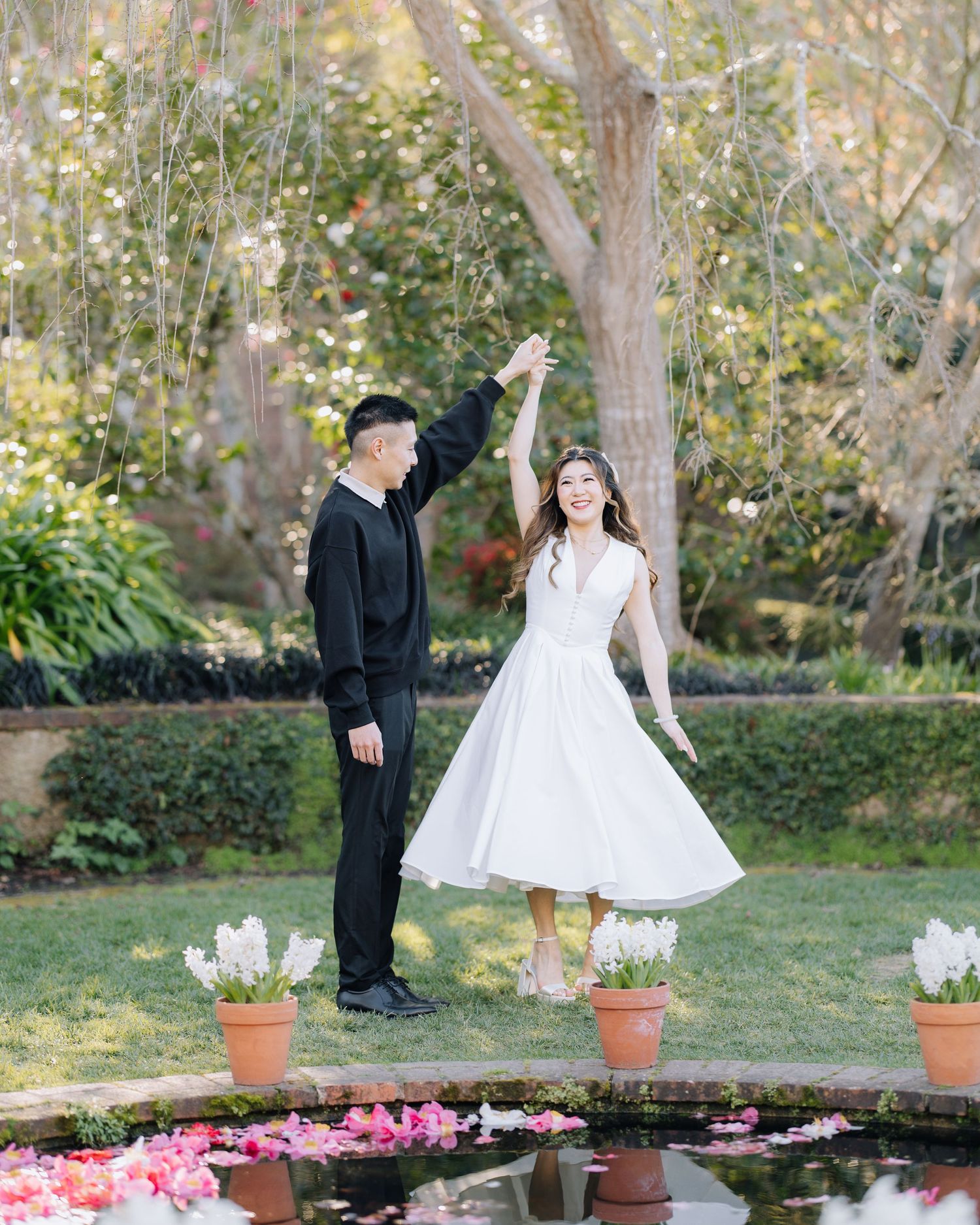 Couple dancing joyfully in a garden with pink petals and blooming spring flowers during Filoli engagement photoshoot.