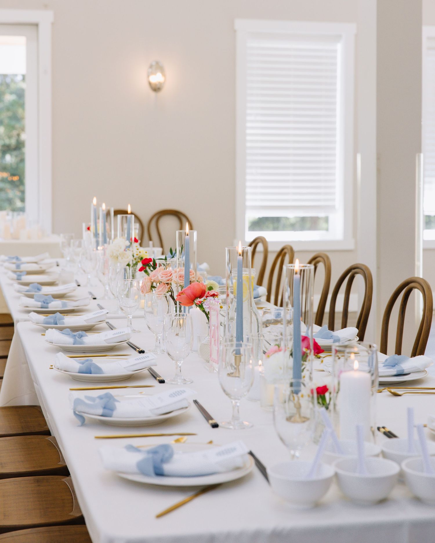 Elegant long reception table with blue taper candles, gold flatware, white linens, and bentwood chairs.