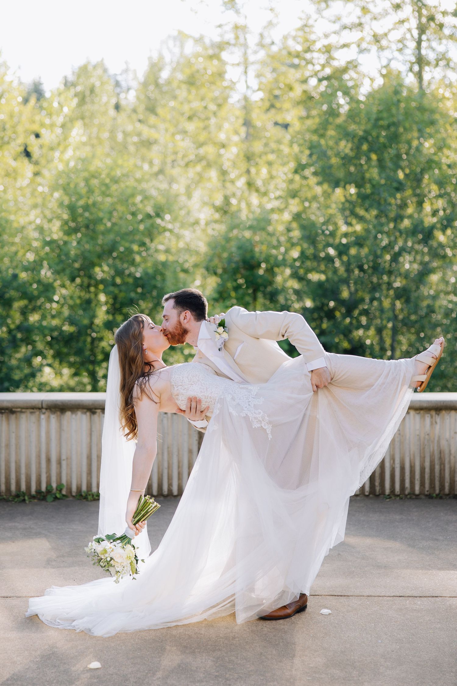 Groom lifts bride in white lace gown for a kiss outdoors with lush green trees in background in Seattle wedding ceremony.