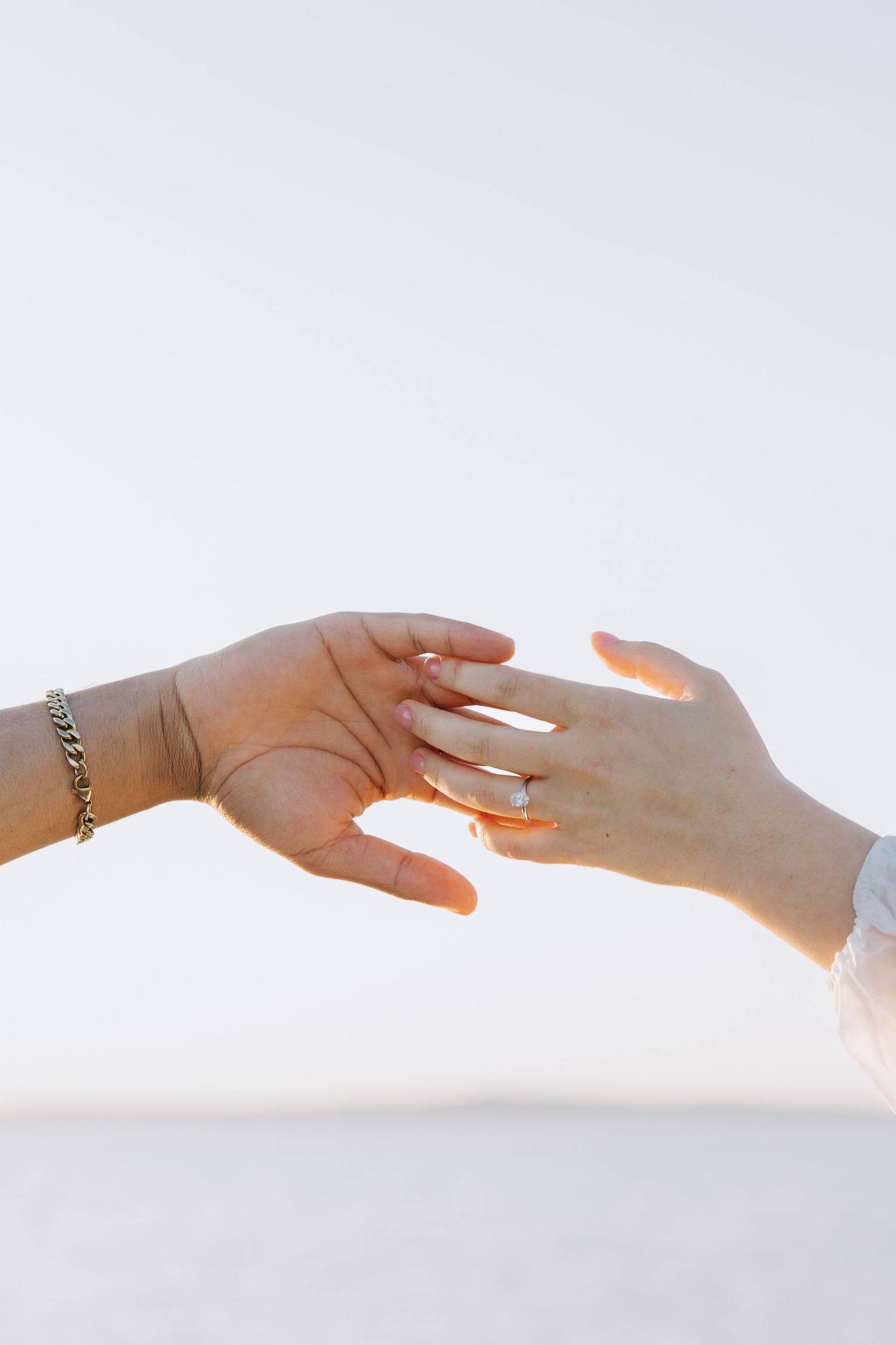 Close-up of couple's hands touching, woman wearing engagement ring, man with silver chain bracelet during beach engagement photoshoot.