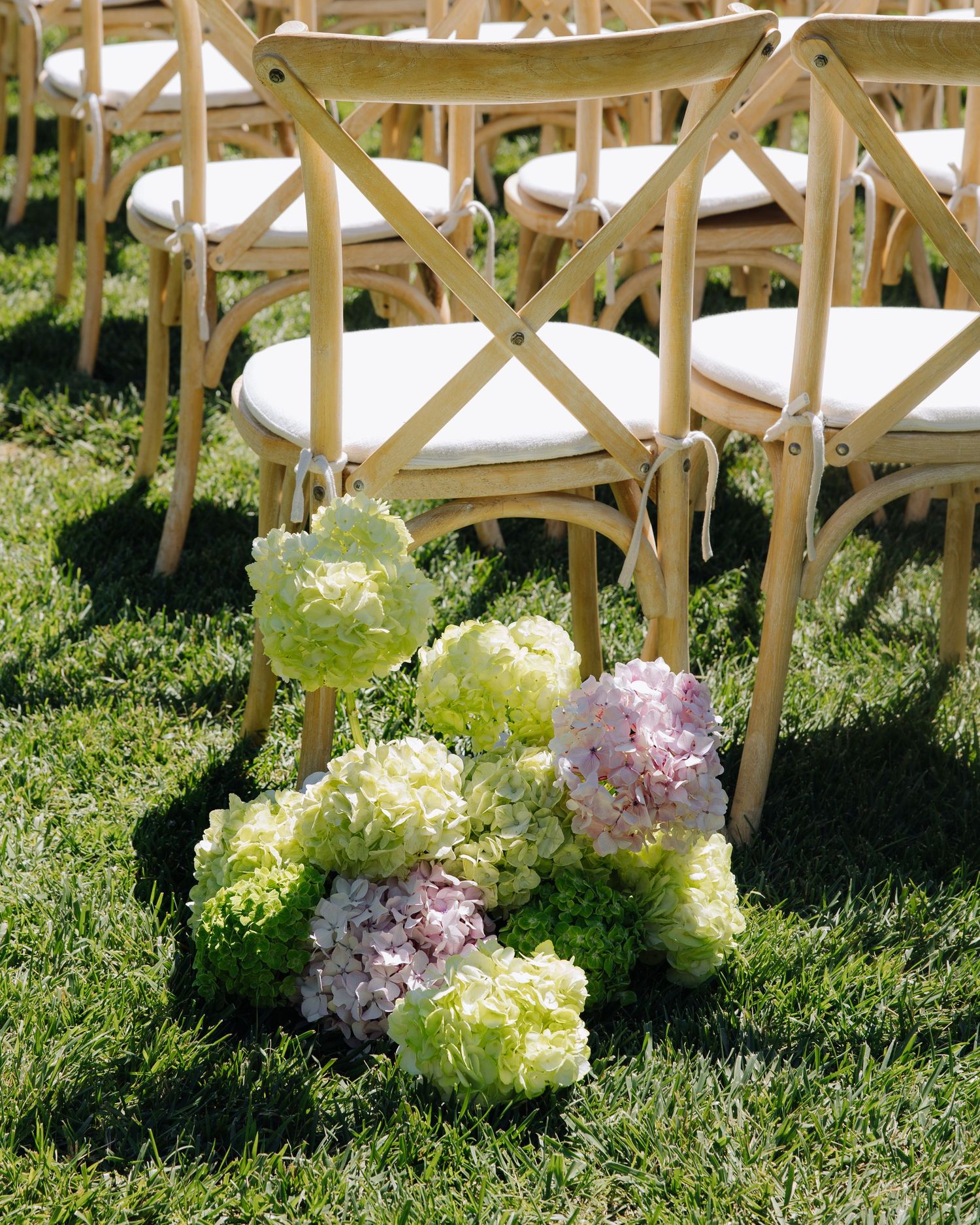 Green and pink hydrangea floral arrangements placed along grass aisle beside wooden cross-back chairs.