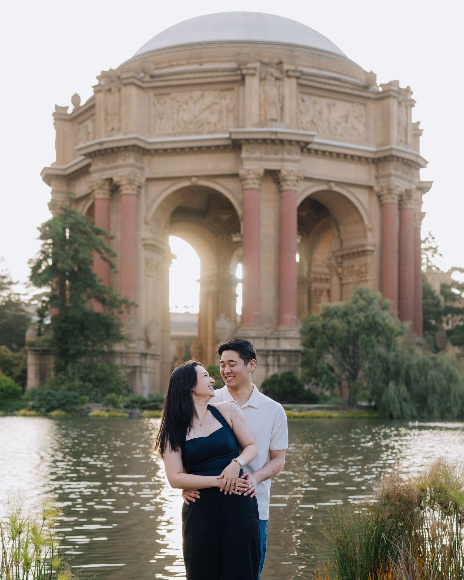 Couple embraces in front of the Palace of Fine Arts rotunda reflected in a lagoon at sunset. Proposal photoshoot in San Francisco.