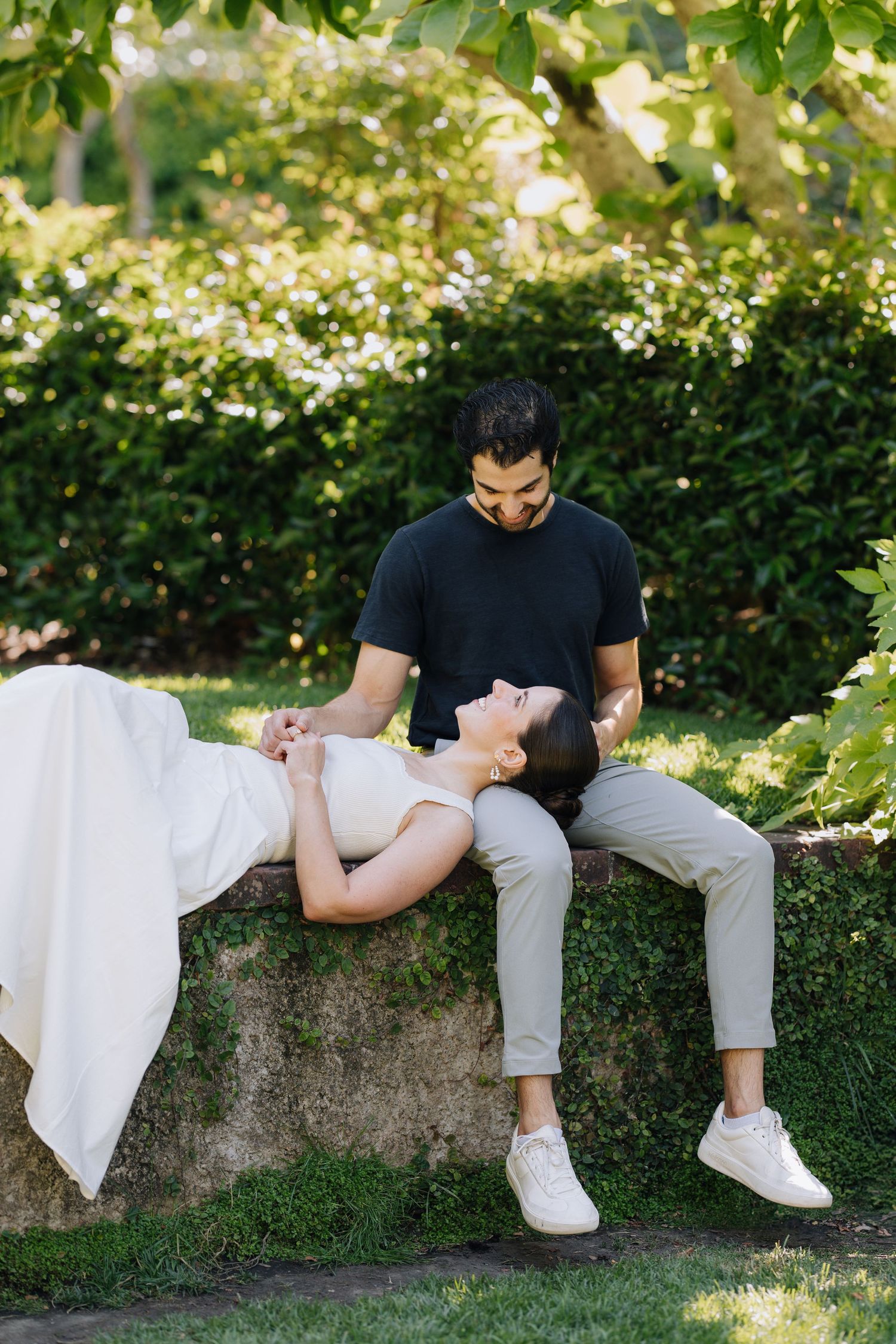 Couple relaxing on stone bench in green garden, woman lying back laughing joyfully during Filoli engagement photoshoot.