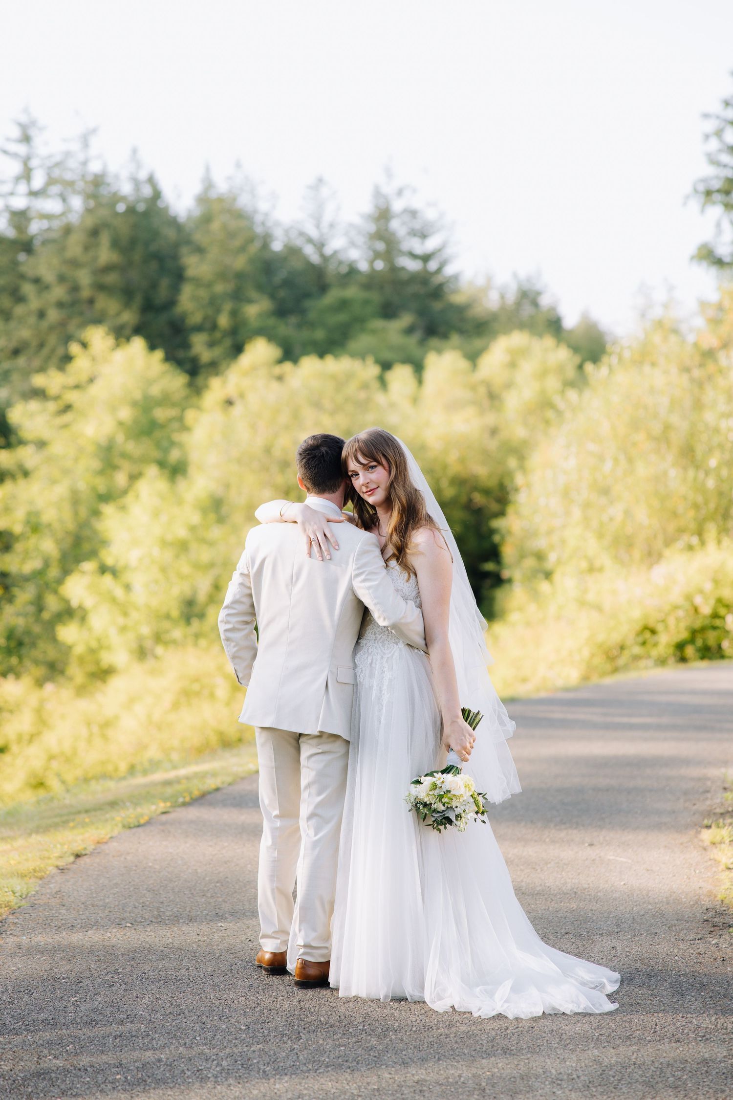 Bride and groom embracing on a scenic tree-lined path, bride holding a white bouquet.