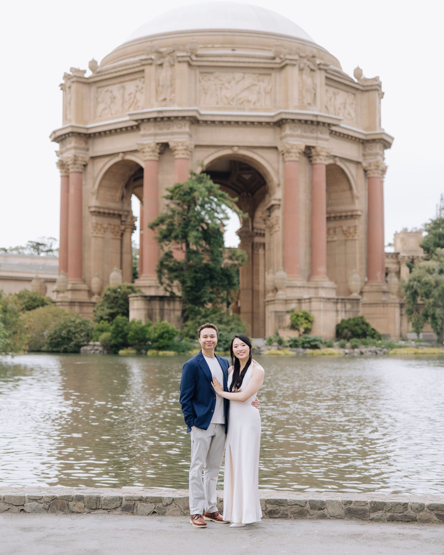 Smiling couple posing together by the Palace of Fine Arts lagoon with the rotunda in background during engagement photoshoot.