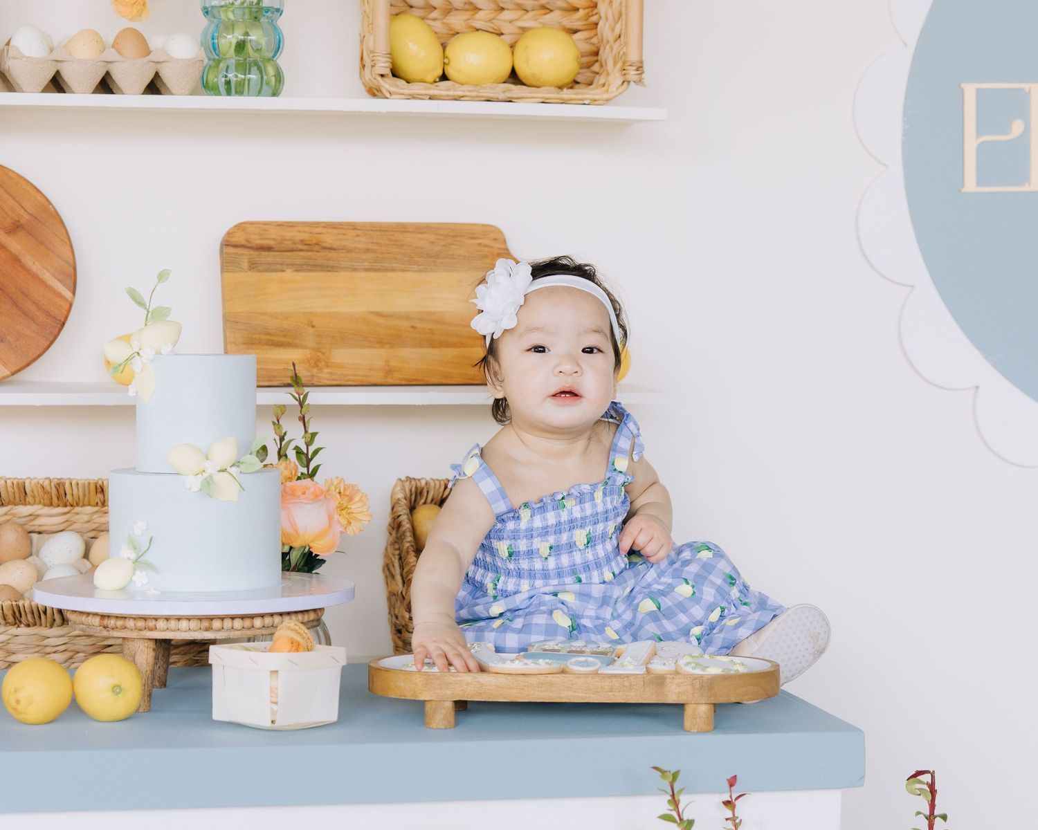 Adorable baby girl in blue gingham dress sitting at decorated table with cake  at baby's first birthday.