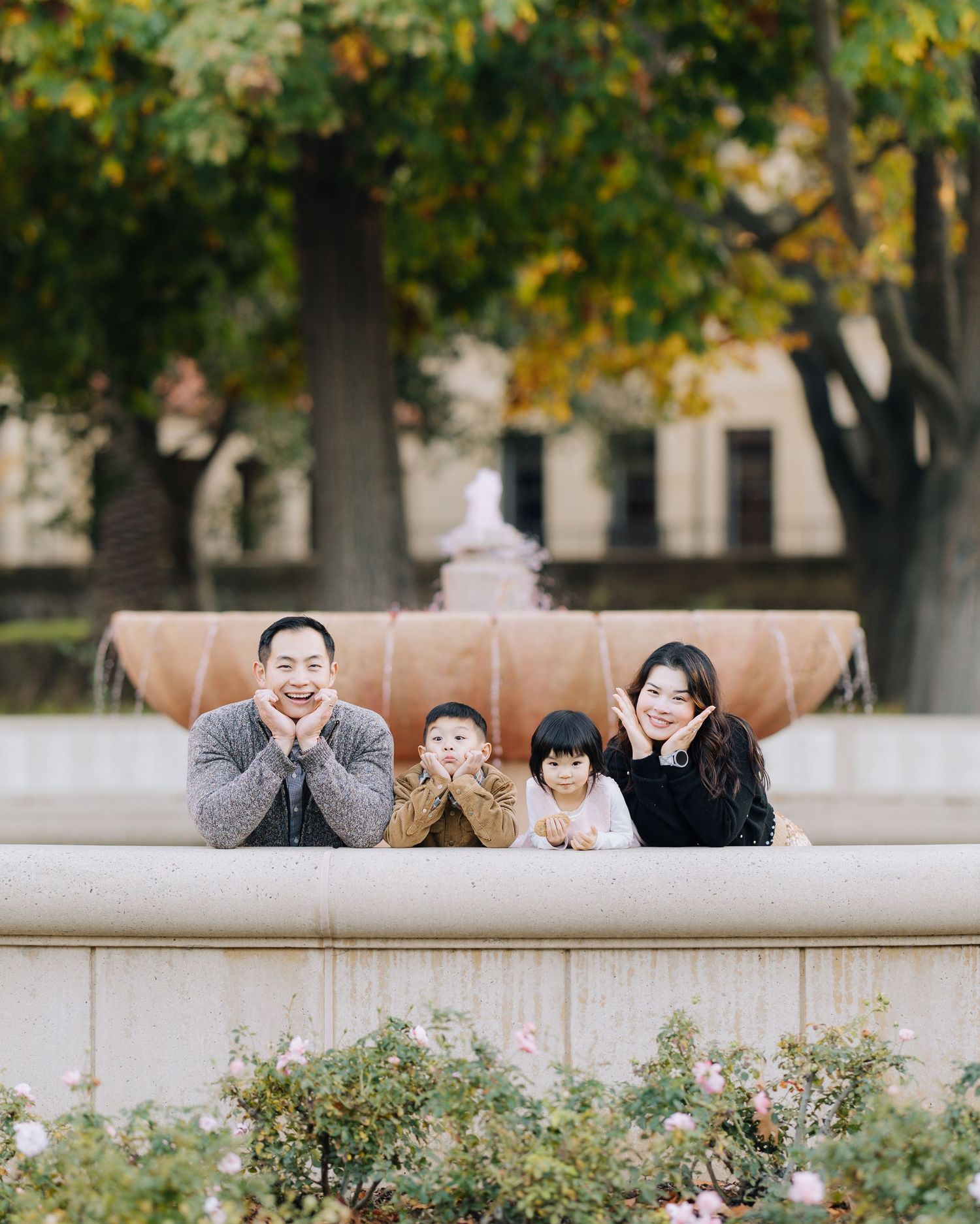 Happy Asian family of four posing playfully at fountain with autumn trees behind at Stanford family photoshoot.