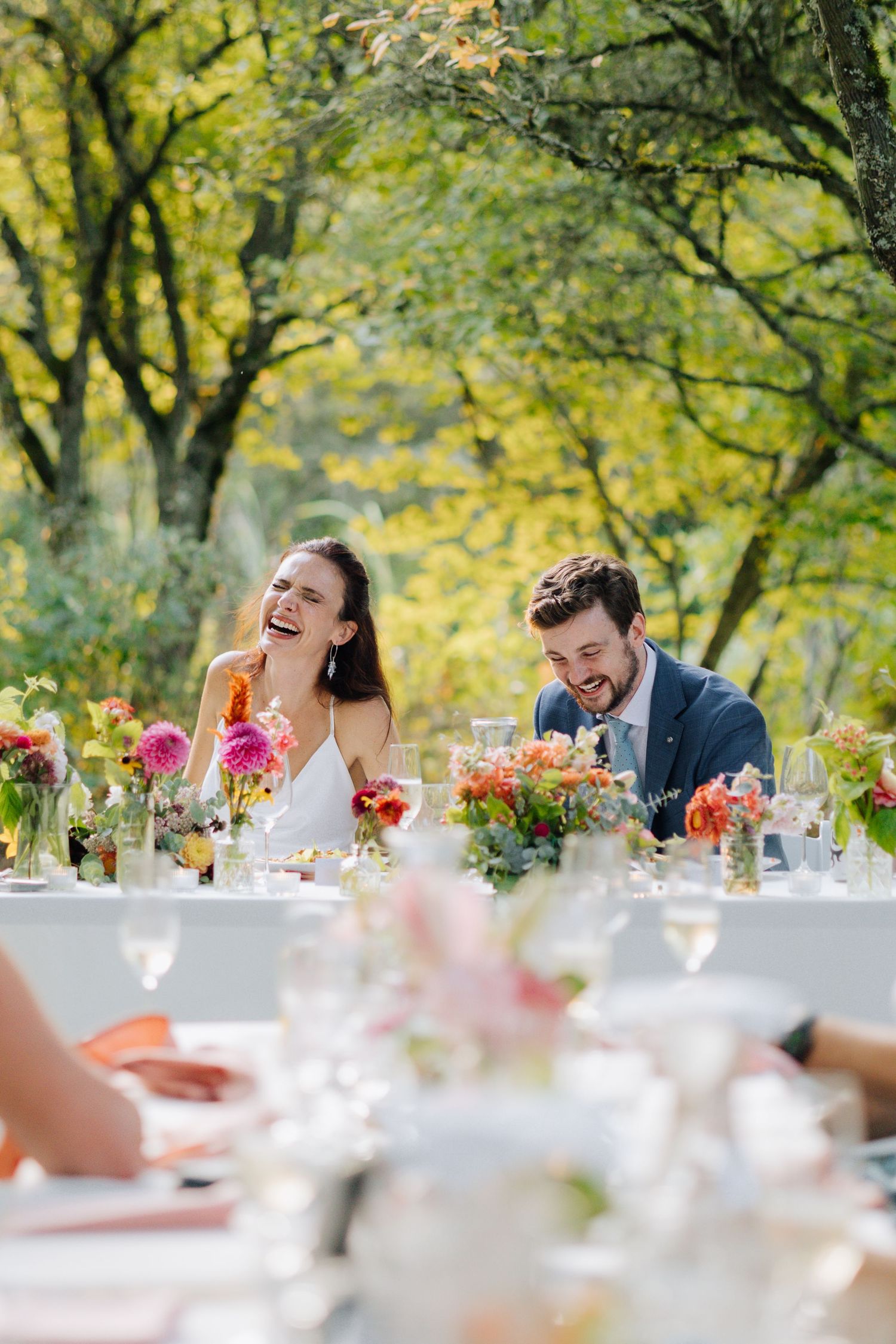 Bride and groom laughing joyfully at outdoor wedding reception table adorned with colorful floral arrangements.