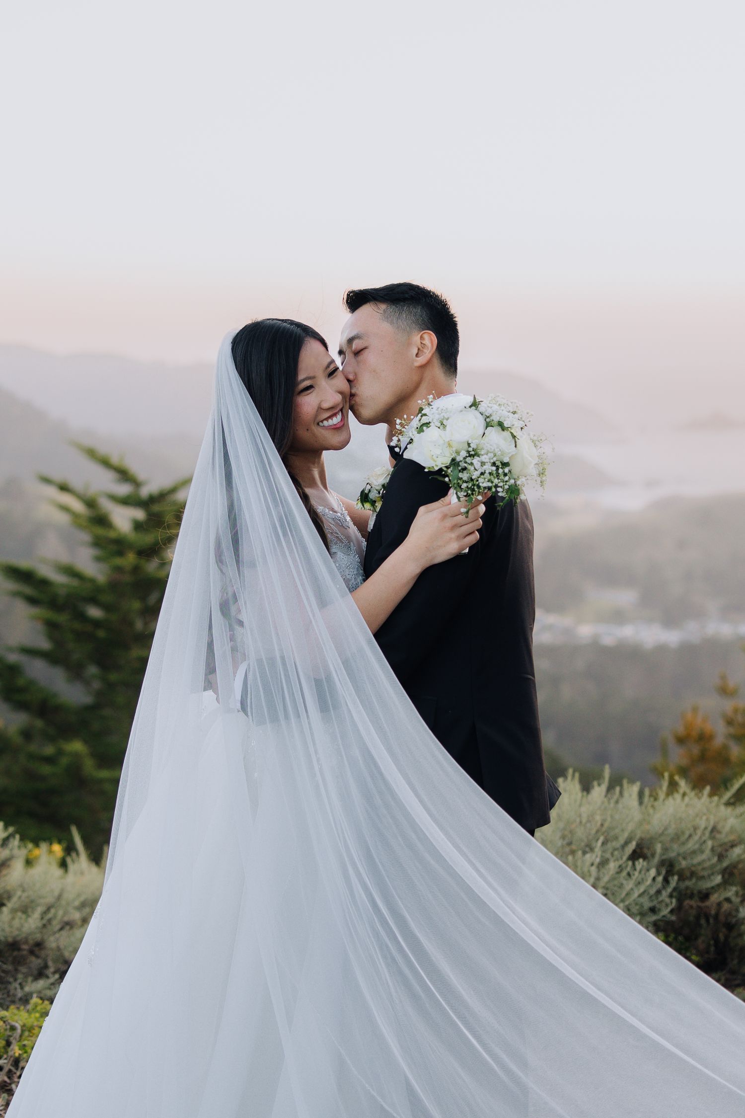 Asian bride and groom share a romantic kiss on a mountain overlook, bride wearing long veil holding white bouquet.