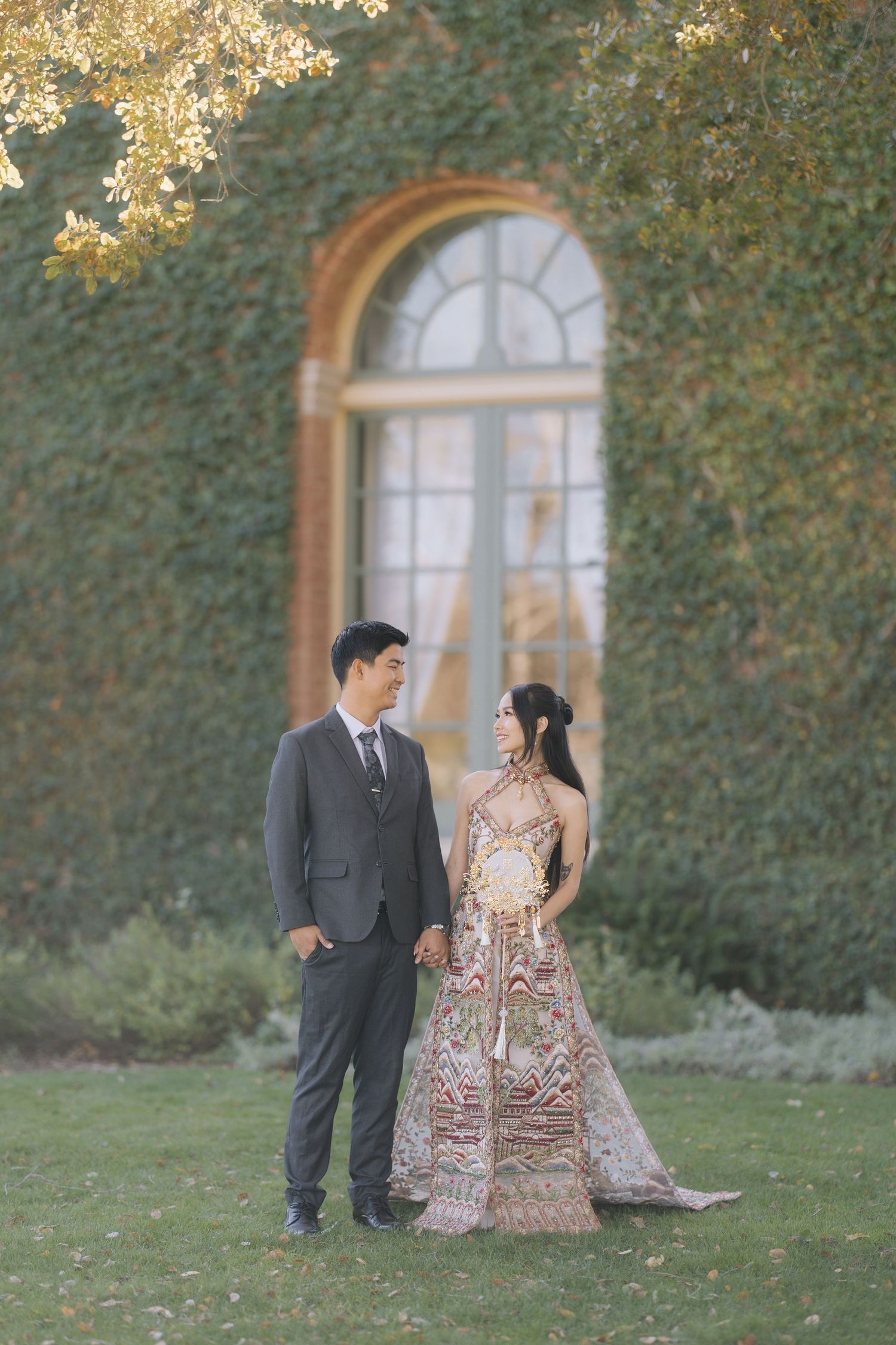 Couple holding hands in garden, woman in ornate embroidered gown, man in gray suit during Filoli wedding photoshoot.
