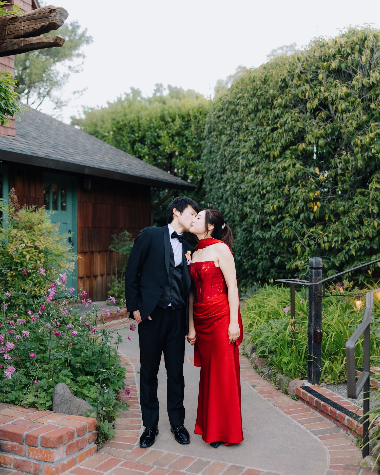 Couple sharing a kiss on a brick pathway, man in black tuxedo and woman in red gown, surrounded by lush garden greenery.