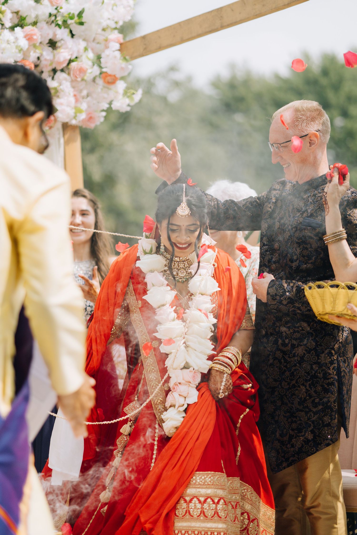 Bride in red saree with floral garland at Indian wedding ceremony with guests throwing rose petals.