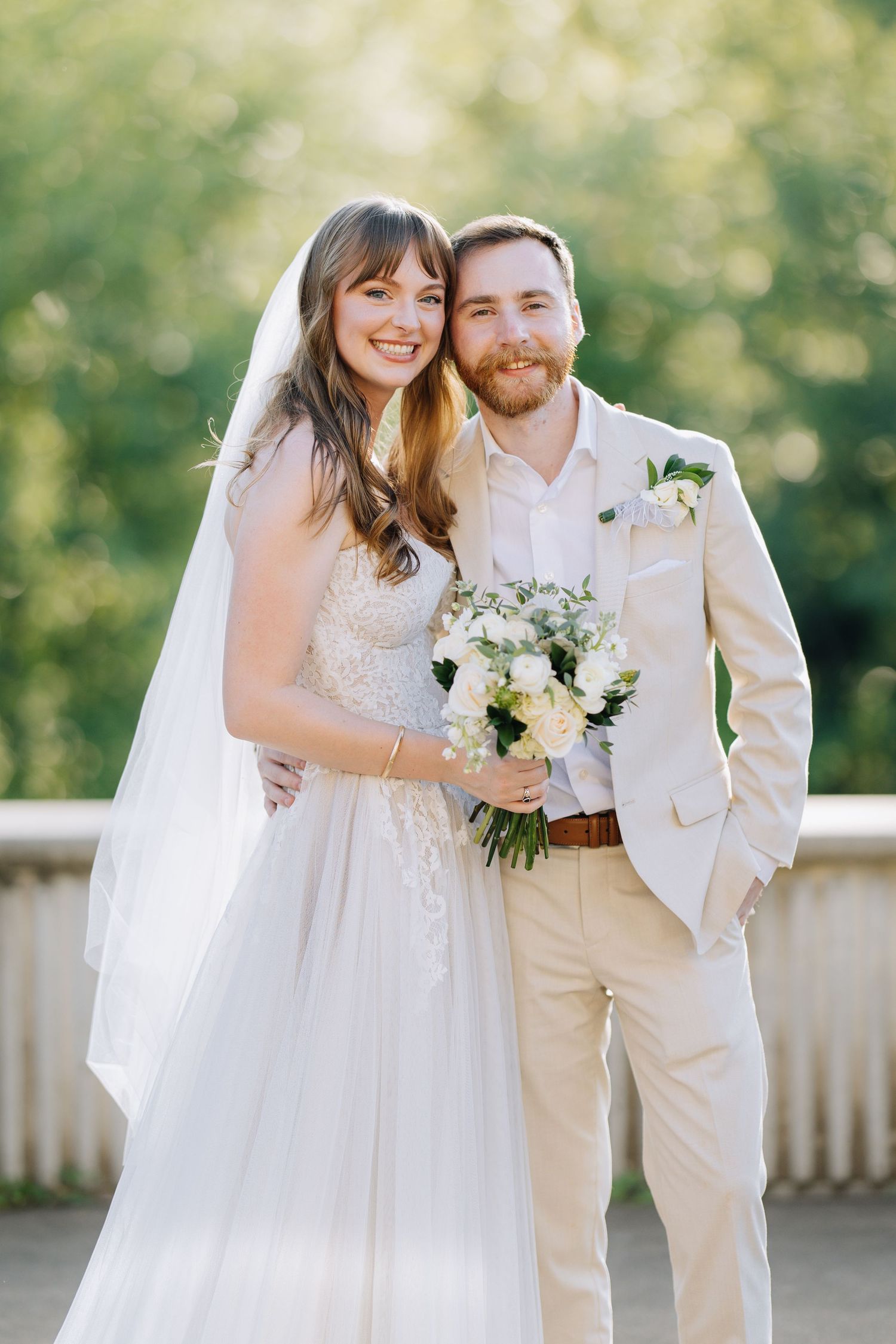 Smiling bride in white lace gown and veil holds bouquet beside groom in beige suit with boutonniere outdoors.