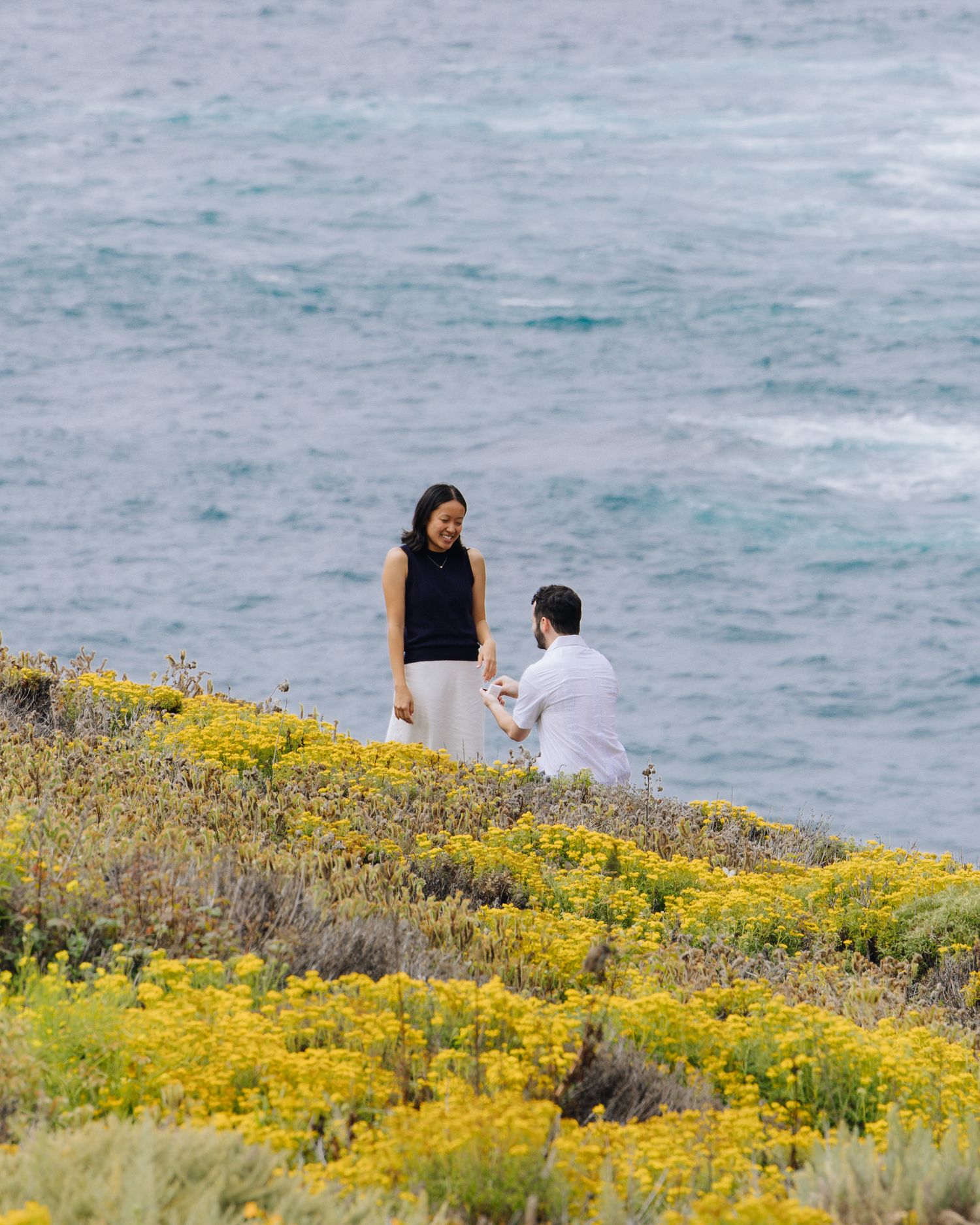 Man proposing on one knee to his partner on a flower-covered coastal hillside during surprise proposal in Carmel.